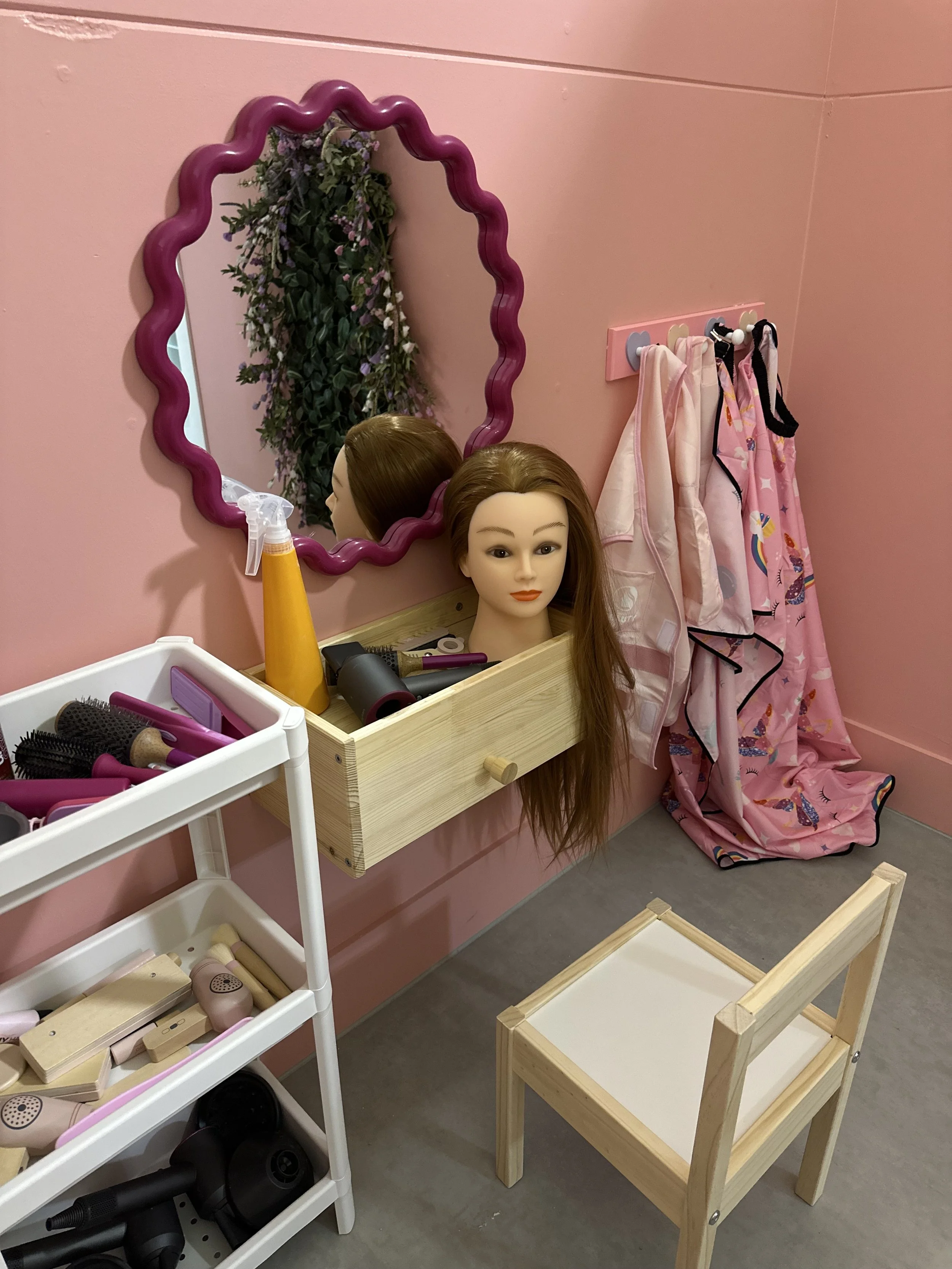 A pink dollhouse dressing room with a mannequin head, mirror, and children's clothing, hairbrushes, and hair styling tools.