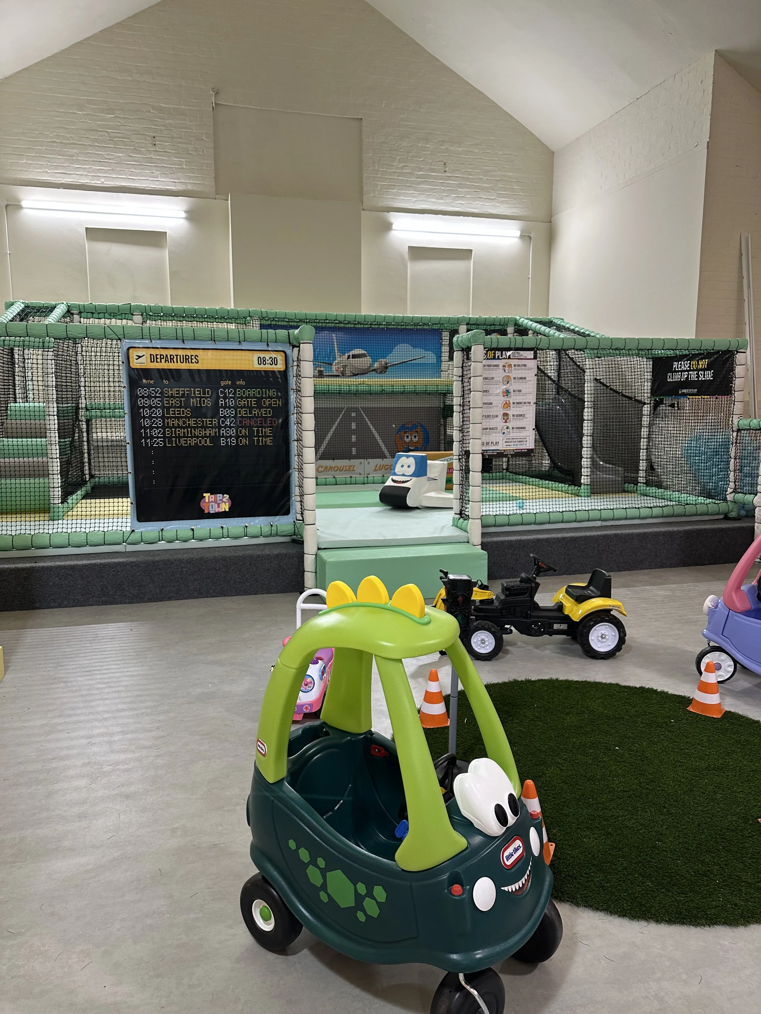 Indoor children's play area with toy cars, a fence around a climbing structure, and a digital departure board. The slide inside the structure has a sign warning about climbing.