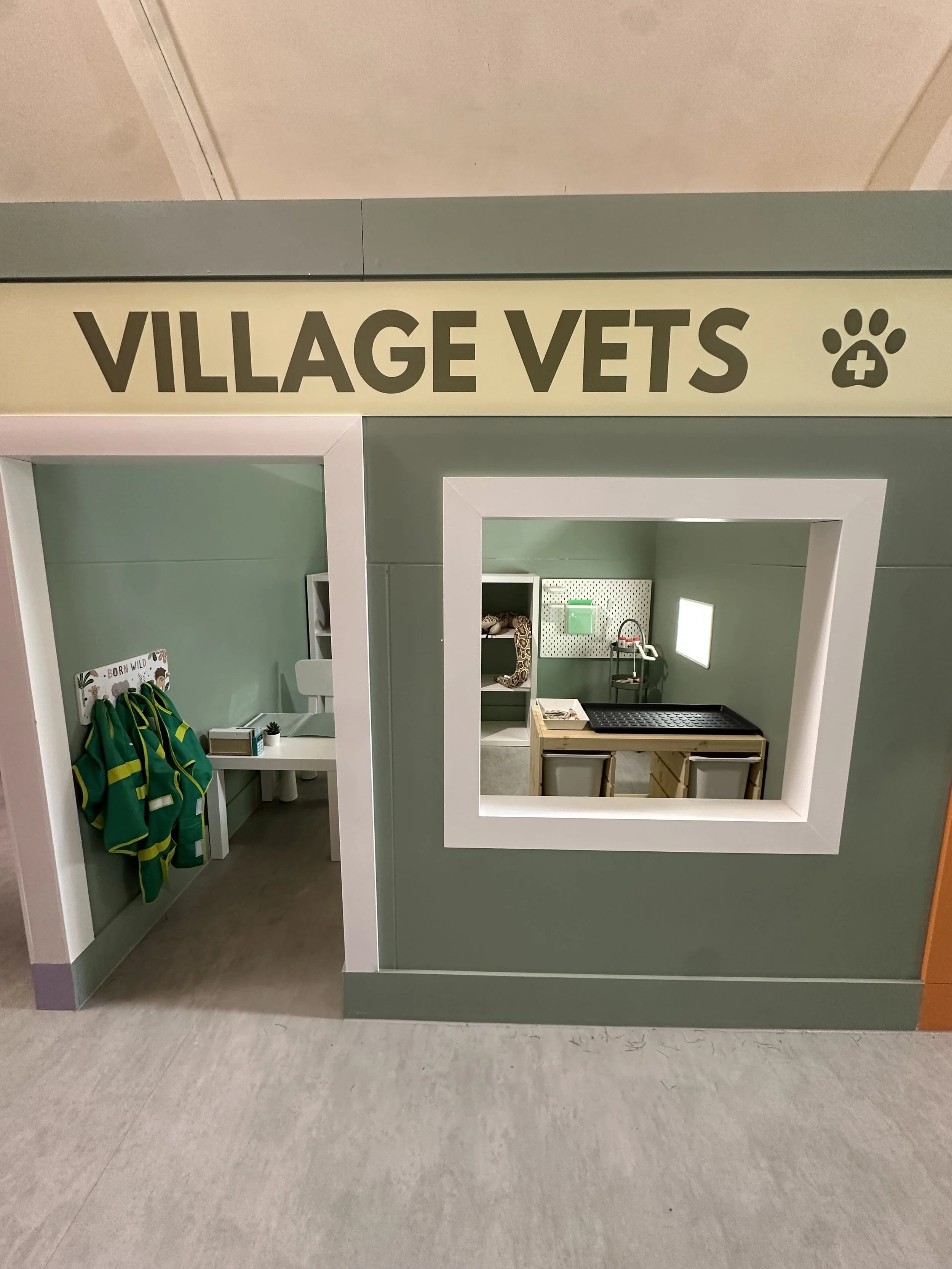 A miniature play area labeled 'VILLAGE VETS' with a small doorway, a window, and furniture including hanging jackets, a sink, and veterinary tools.
