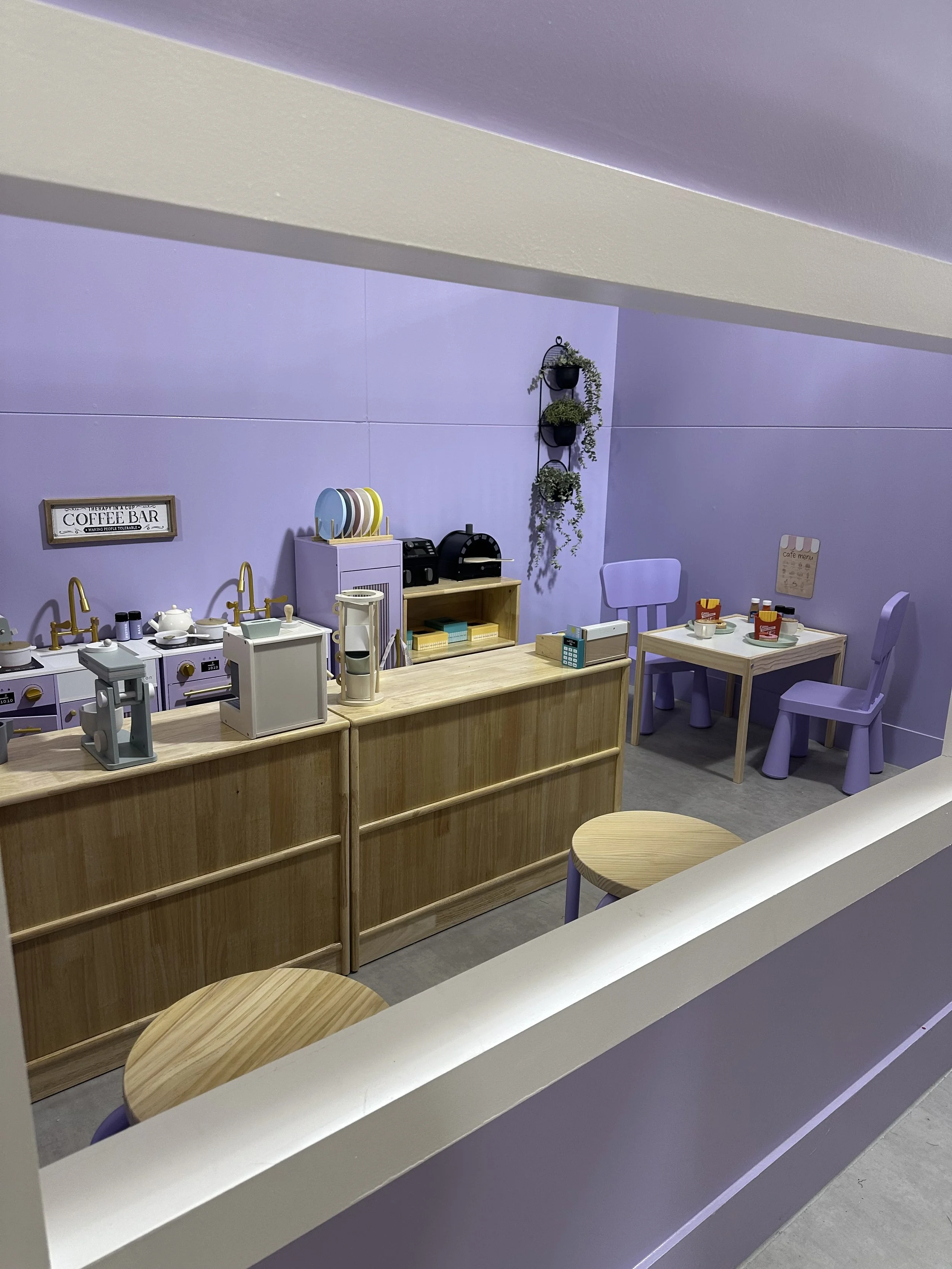 A cheerful play cafe setup with a small counter, wooden tables and chairs, and purple accents including small chairs and wall decor. The counter has toy appliances and dishes, and there are plants on the wall.