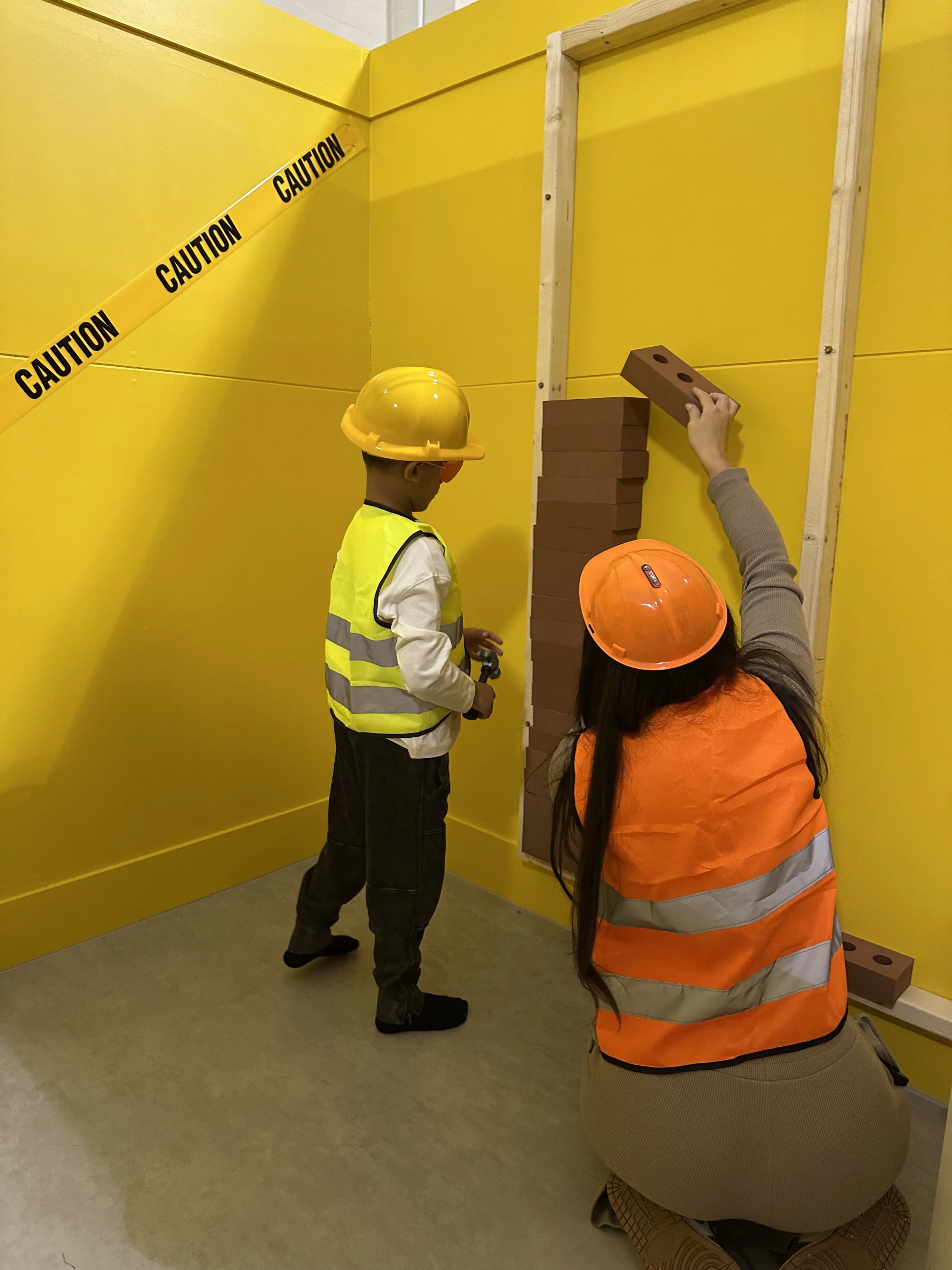 A woman and a boy wearing construction helmets and safety vests building a wall with bricks inside a yellow room, with caution tape on the wall.