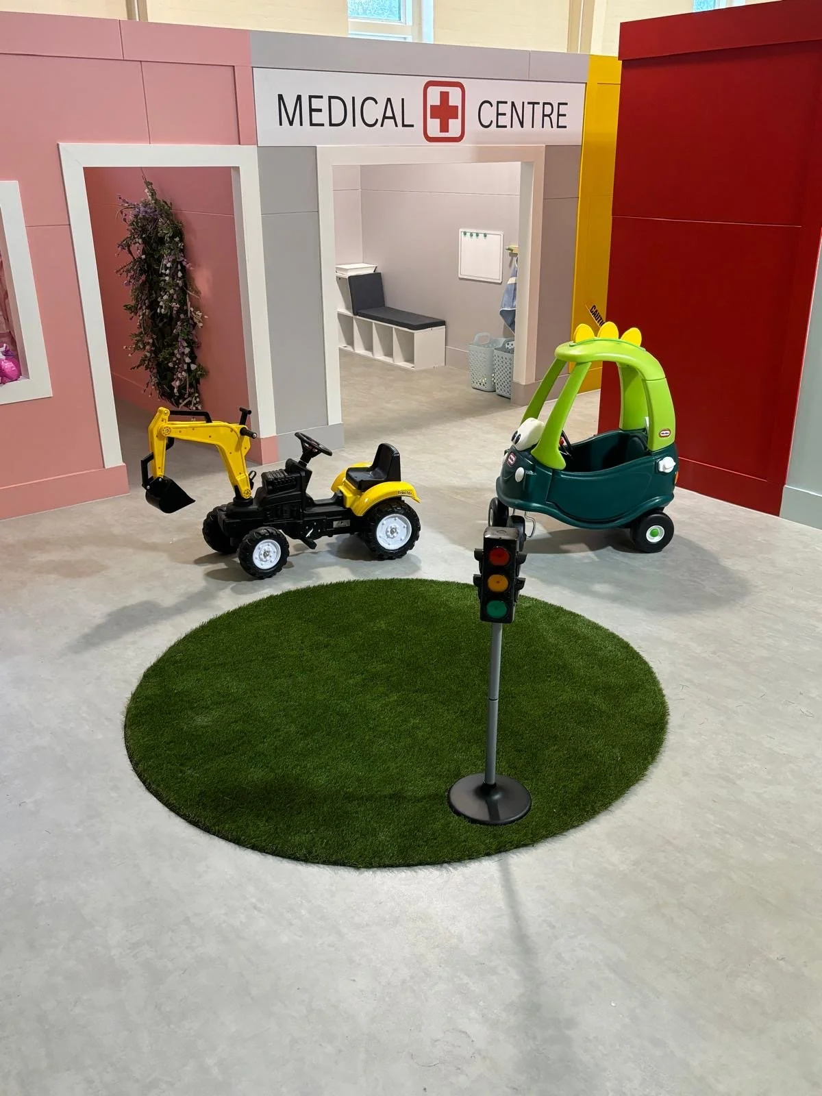 Play area with toy tractor, toy car, traffic light, and green grass circle in front of a mock medical center entrance in a children's play space.