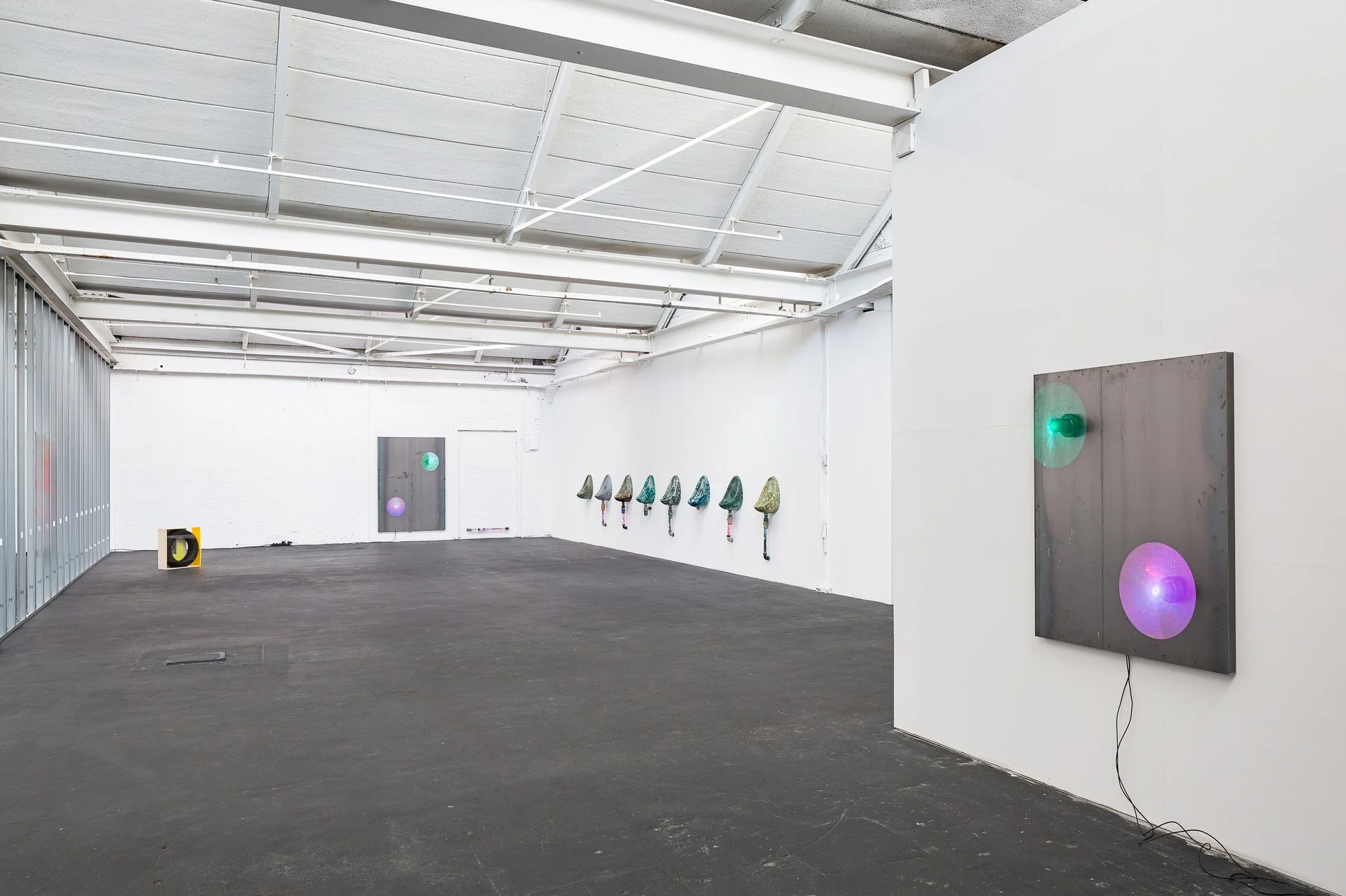 An art gallery with white walls and a black floor displaying several sculptures of bicycles with colorful wheels mounted on the wall, along with a digital art piece on the right side showing two illuminated circles on a metal panel.