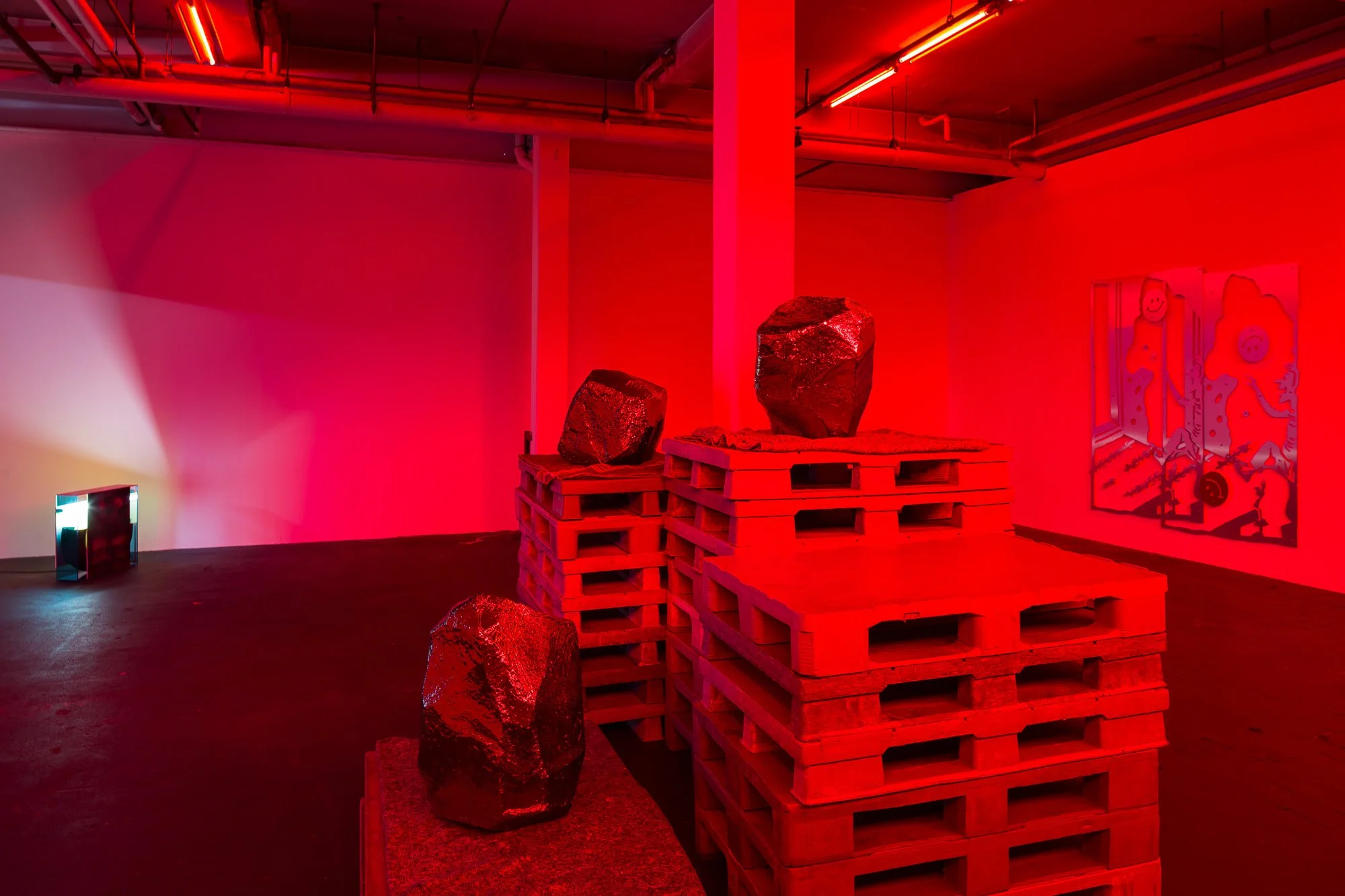 Art installation featuring large dark rocks displayed on wooden pallets in a room illuminated with red light, with a colorful artwork on the wall in the background.