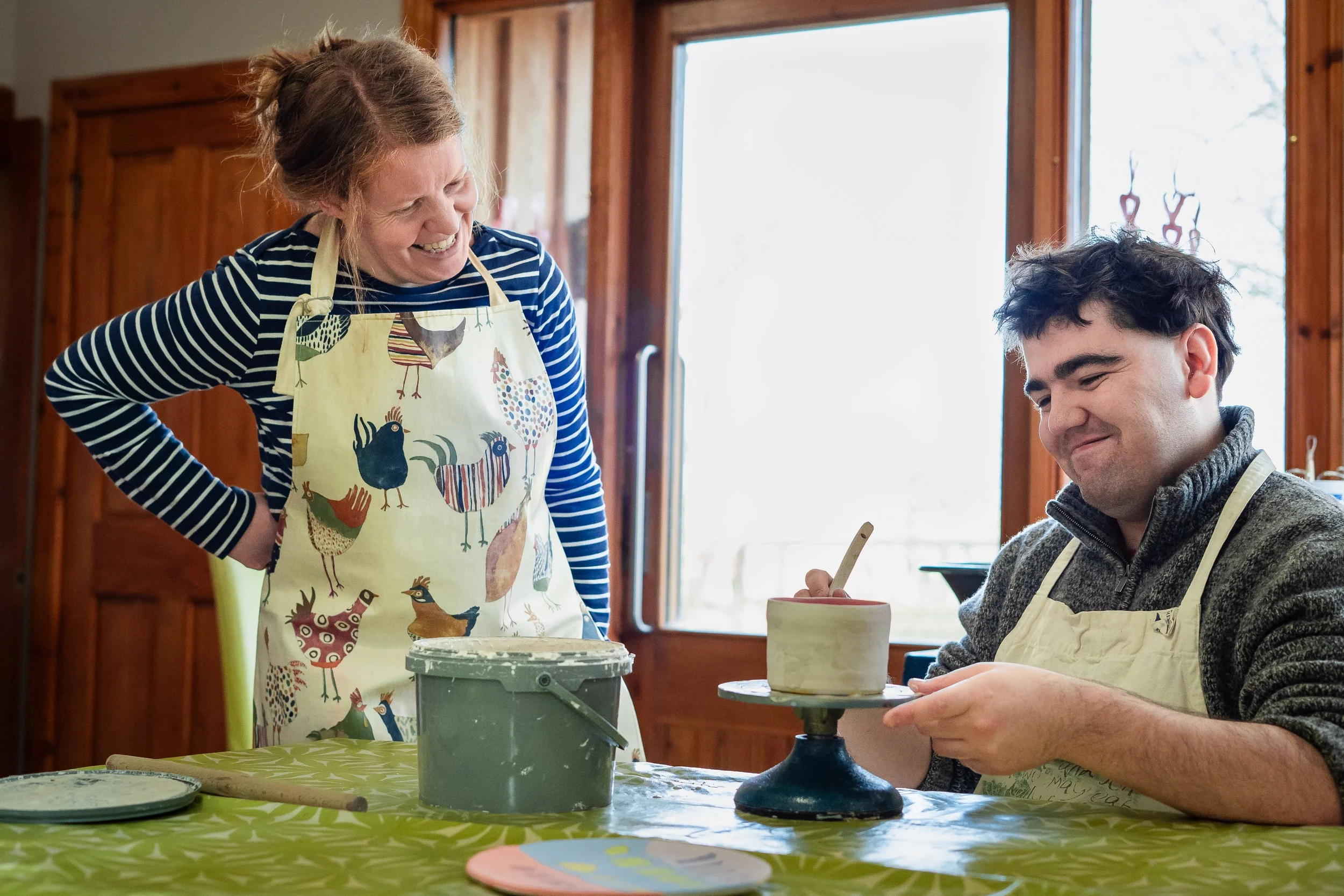 A woman is smiling and leaning over a table, watching a young man who is decorating pottery on a turntable, with a bright window in the background.