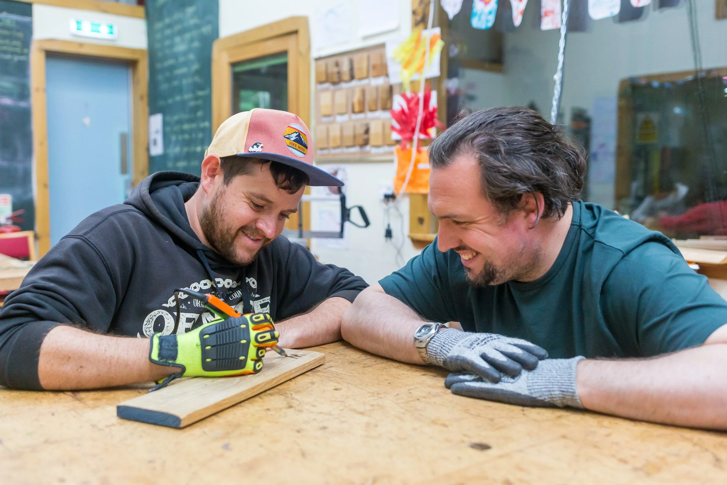 Two men smiling and working together on a woodworking project in a workshop, with one of them holding a pencil and marking a piece of wood, both wearing gloves.