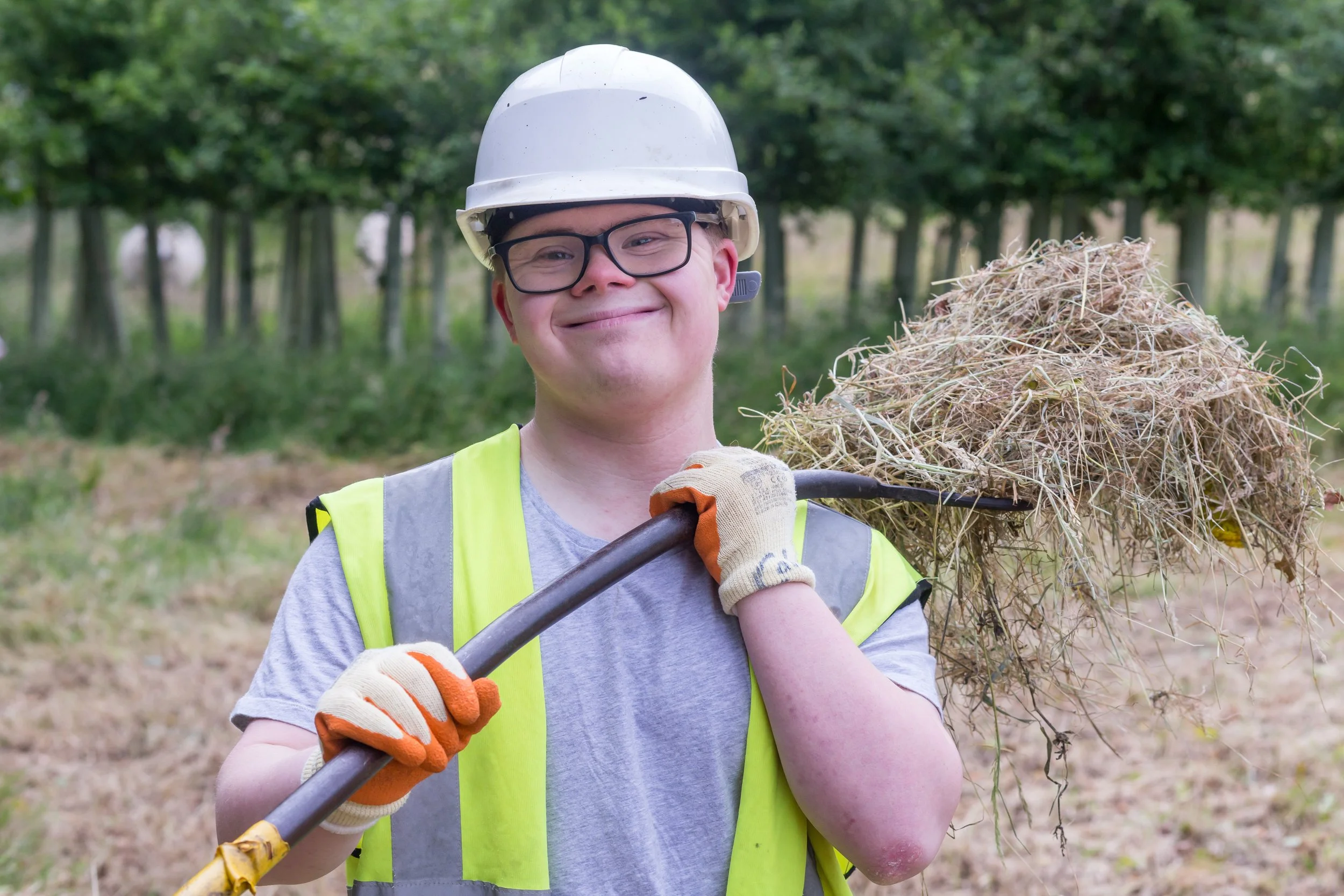 A smiling young man in safety gear, including a white helmet, safety glasses, and a yellow safety vest, holding a rake with a pile of dried grass and weeds on it in a wooded outdoor area.