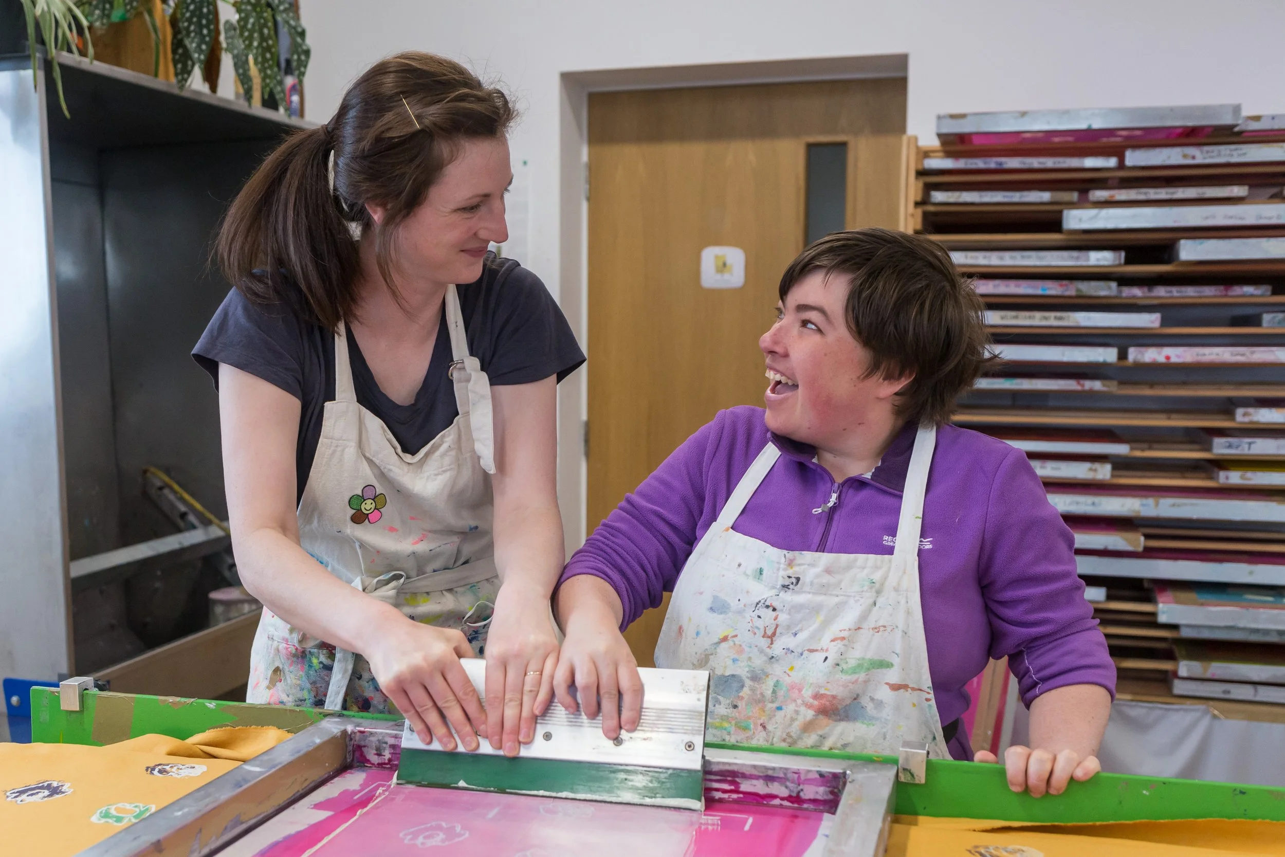 A woman and a boy are working together at a table, using a screen printing machine. They are smiling and looking at each other, wearing aprons covered in paint, in an art room with colorful papers and art supplies.