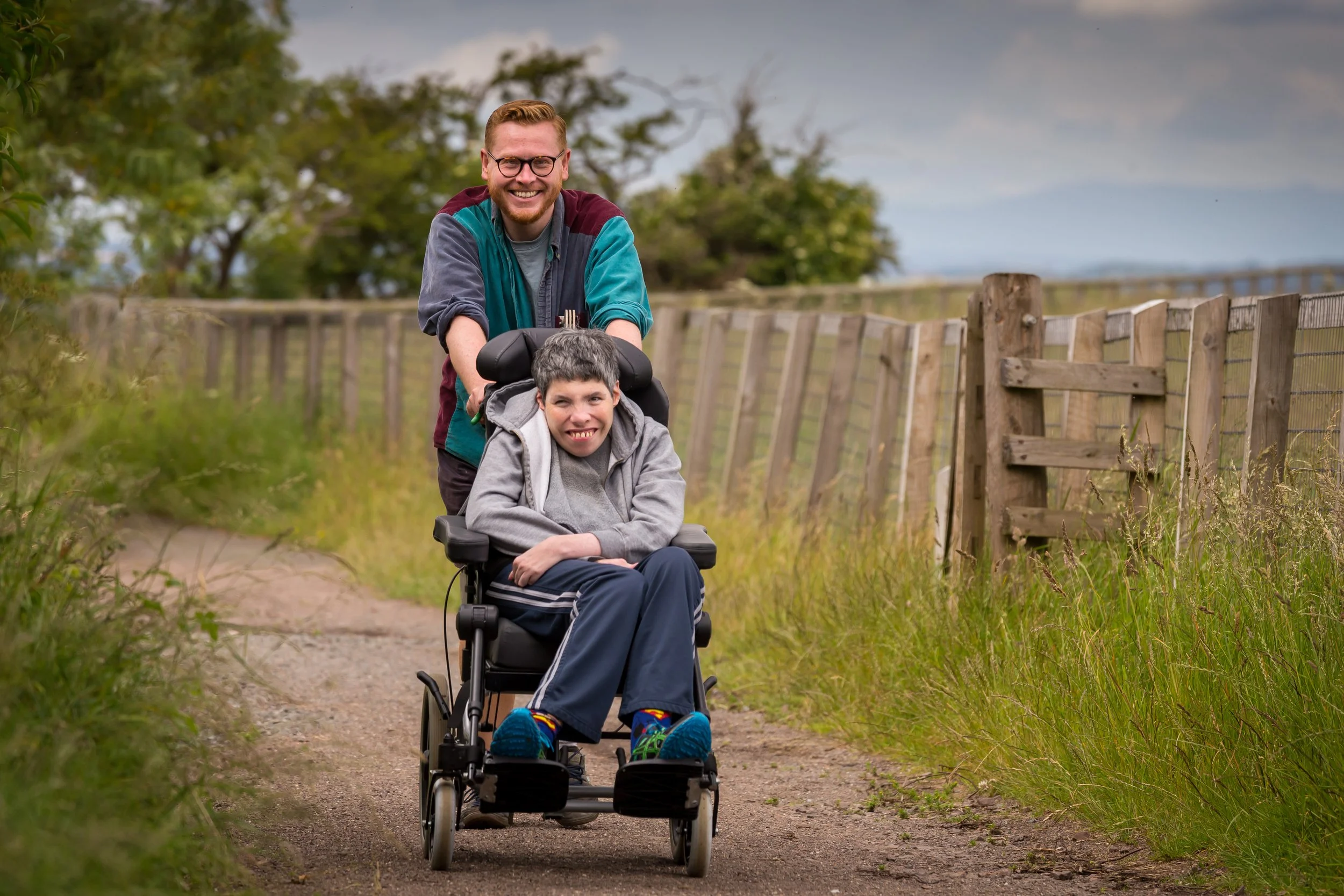 A man and a young person in a wheelchair are outdoors on a dirt path, smiling and enjoying a walk, with a wooden fence and trees in the background.