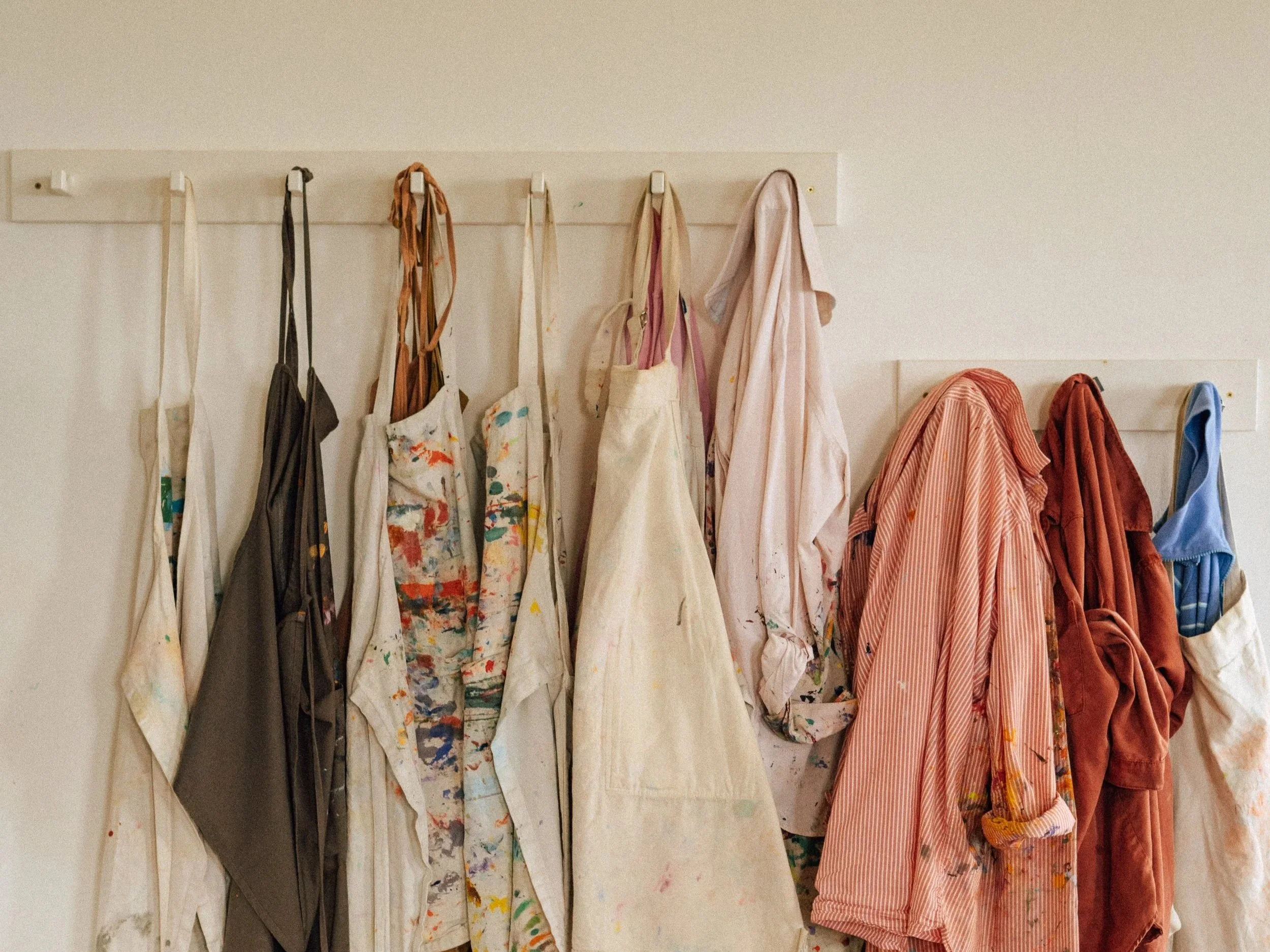 Paint-stained aprons and clothes hanging on hooks on a white wall.