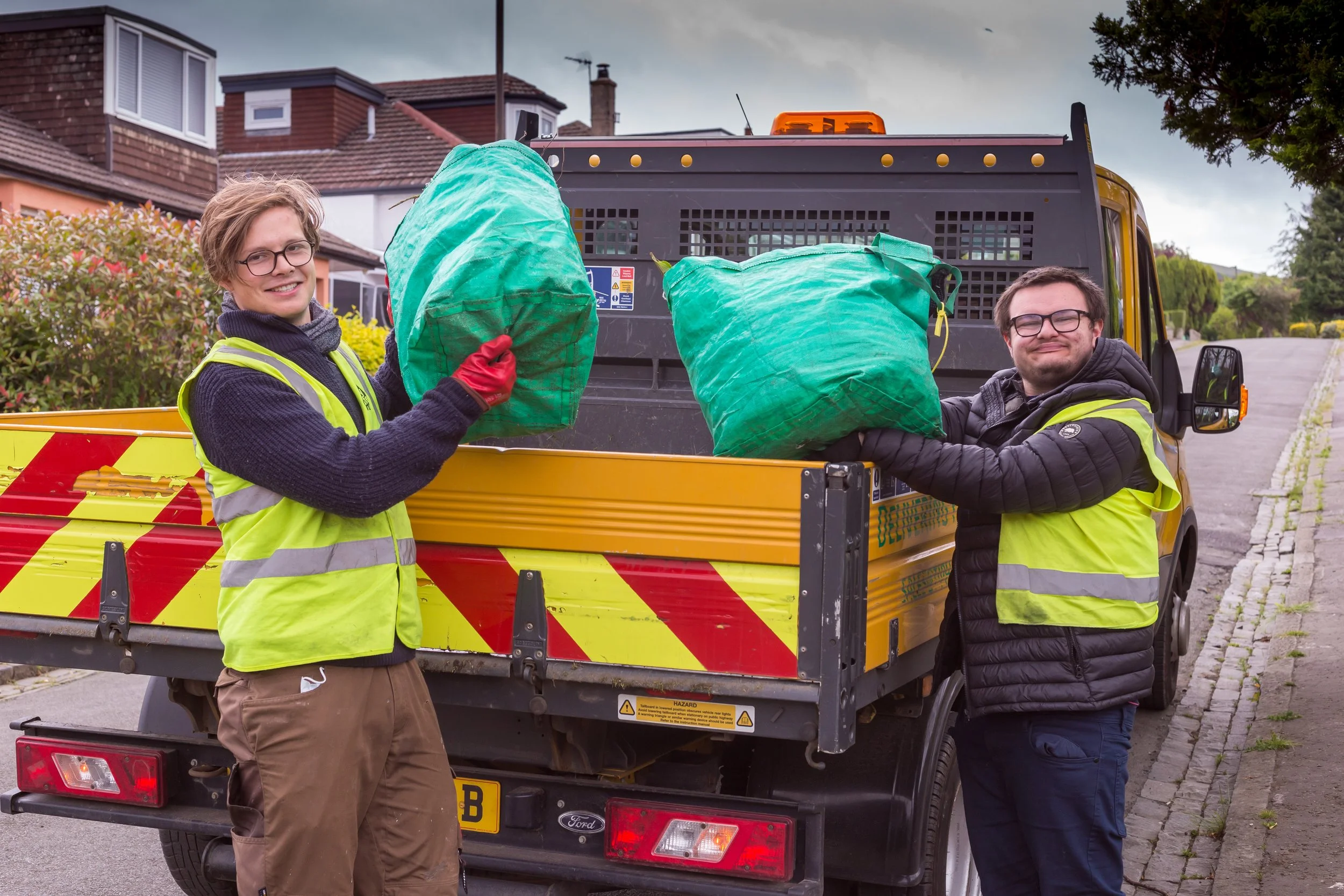 Two workers in high-visibility vests unload large green bags from a yellow work truck parked on a residential street.