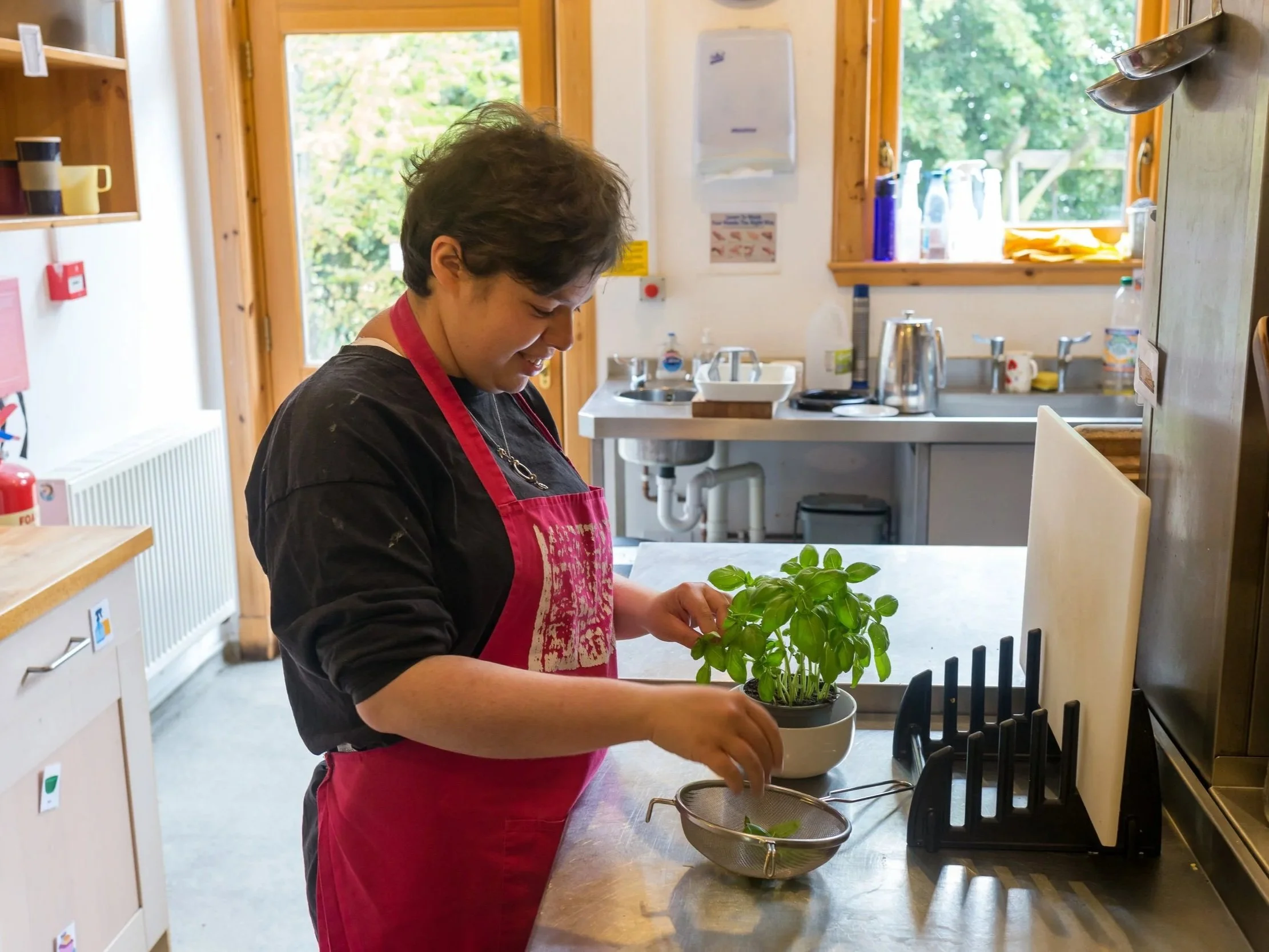 A woman wearing a black shirt and a red apron is preparing herbs in a kitchen, standing at a wooden countertop with a potted basil plant and a sieve.