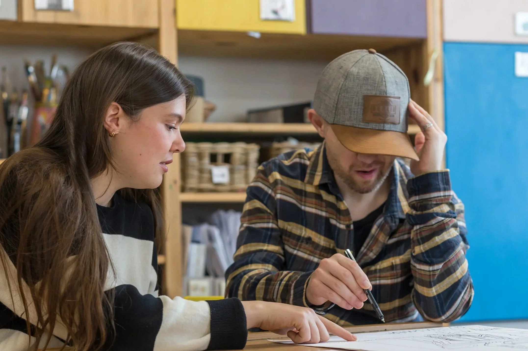 A young woman and a man sit at a table, looking at a document. The woman points at the document, and the man wears a gray cap and a plaid shirt.