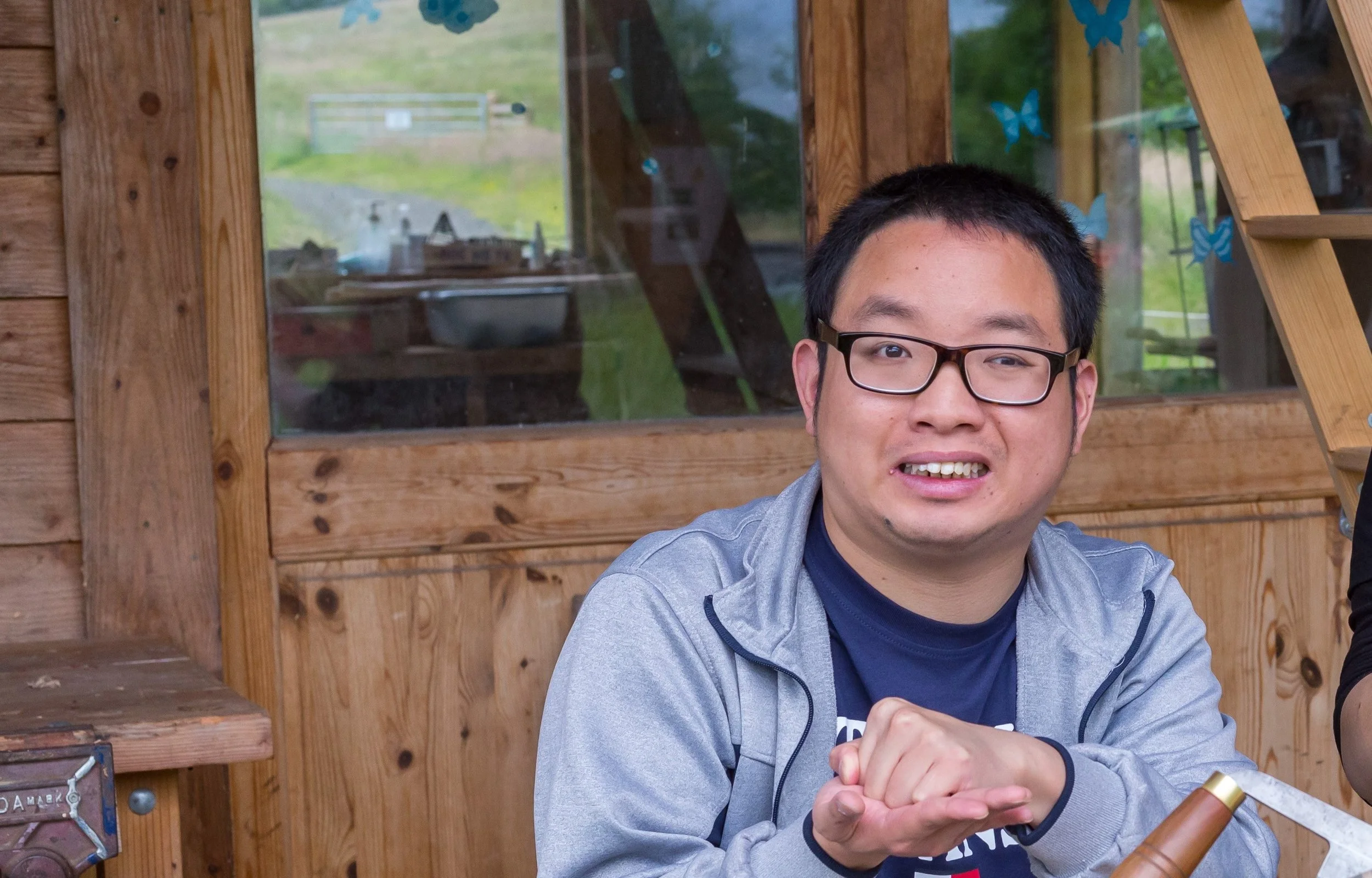 A man with black hair, glasses, and a gray jacket, using sign language and smiling at the camera while sitting at a wooden table inside a rustic cabin.