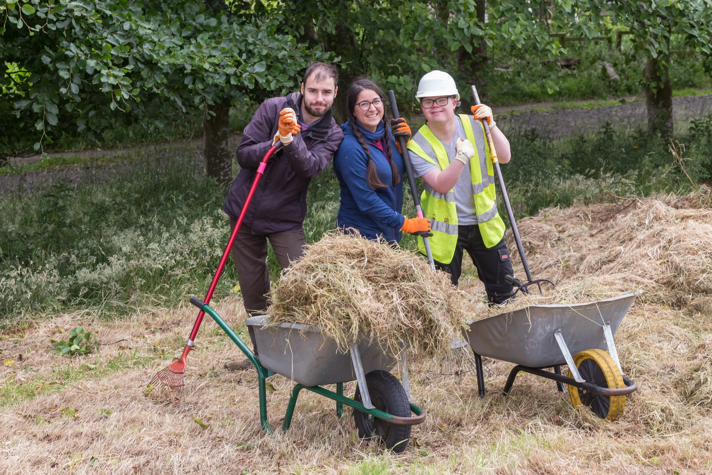 Three people standing outdoors with wheelbarrows filled with hay, smiling and holding gardening tools, with trees and grass in the background.