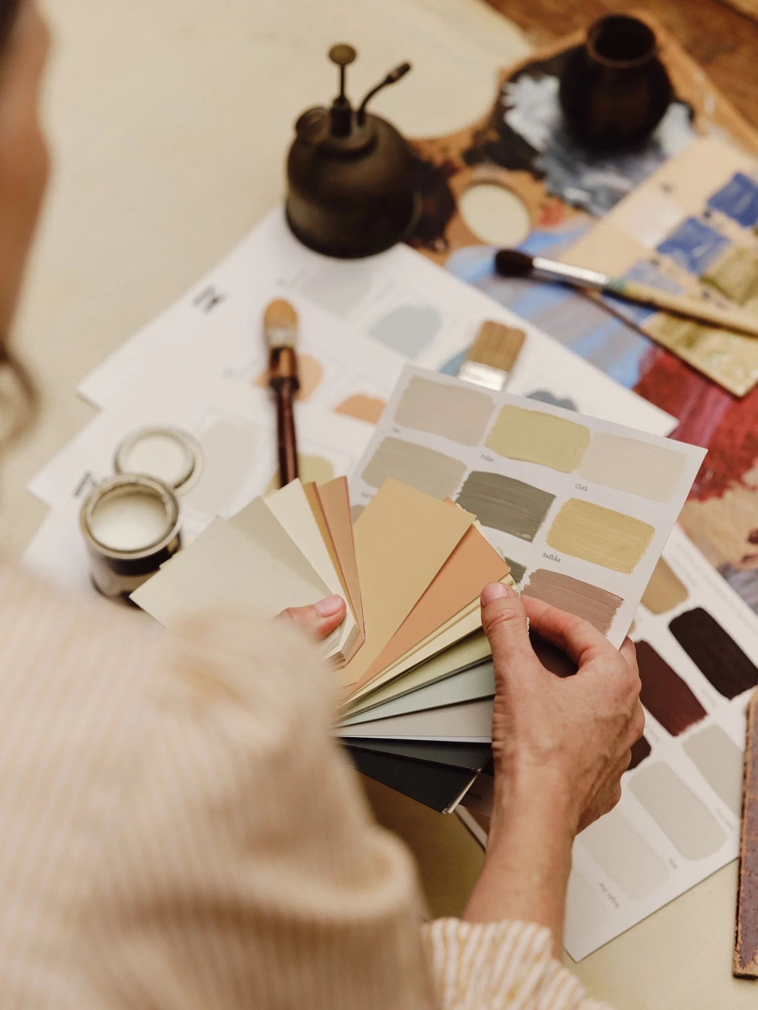 Person holding a fan deck of paint color swatches over a table with paint sample sheets, brushes, tape, and small jars of paint.