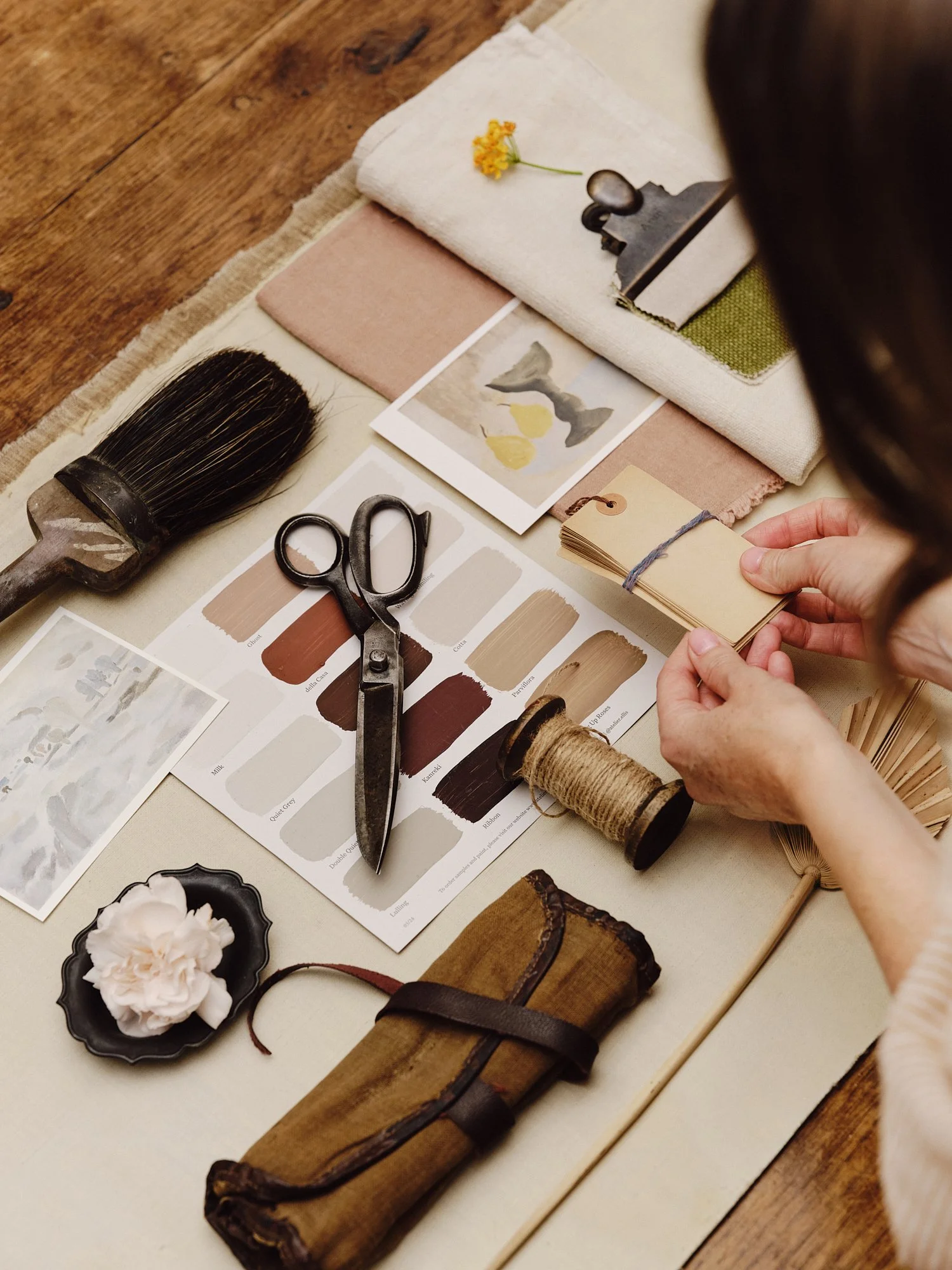 A person working on a creative project with paint swatches, scissors, a paintbrush, swatches of fabric, small photographs, a spool of twine, a rolled-up piece of wood, and a black dish holding a white object. The workspace is on a beige cloth on a wooden surface.
