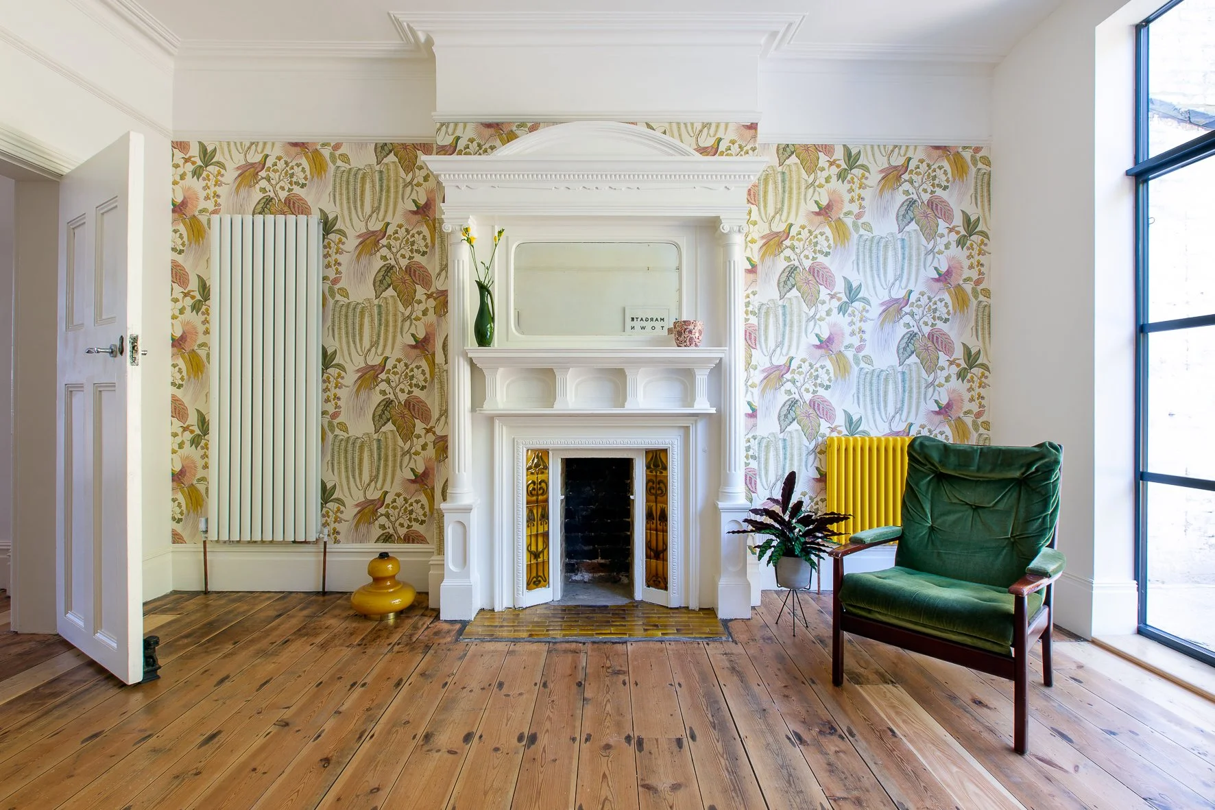 A cozy living room corner with a white ornate fireplace, floral wallpaper, a green armchair, a large window, and decorative items including a vase with flowers, a small plant, and a yellow vase. The hardwood floor adds warmth to the space.
