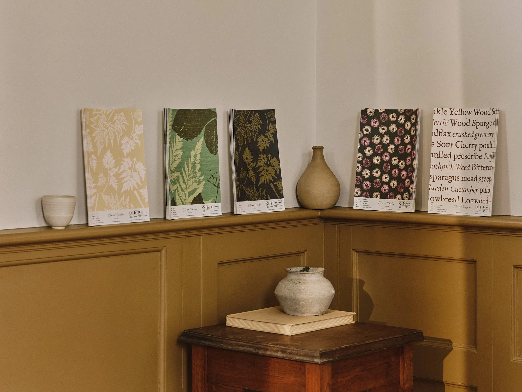 Display of garden-themed notebooks with botanical patterns, small pottery vases, and an earthenware pot on a wooden table against a white and tan wall.