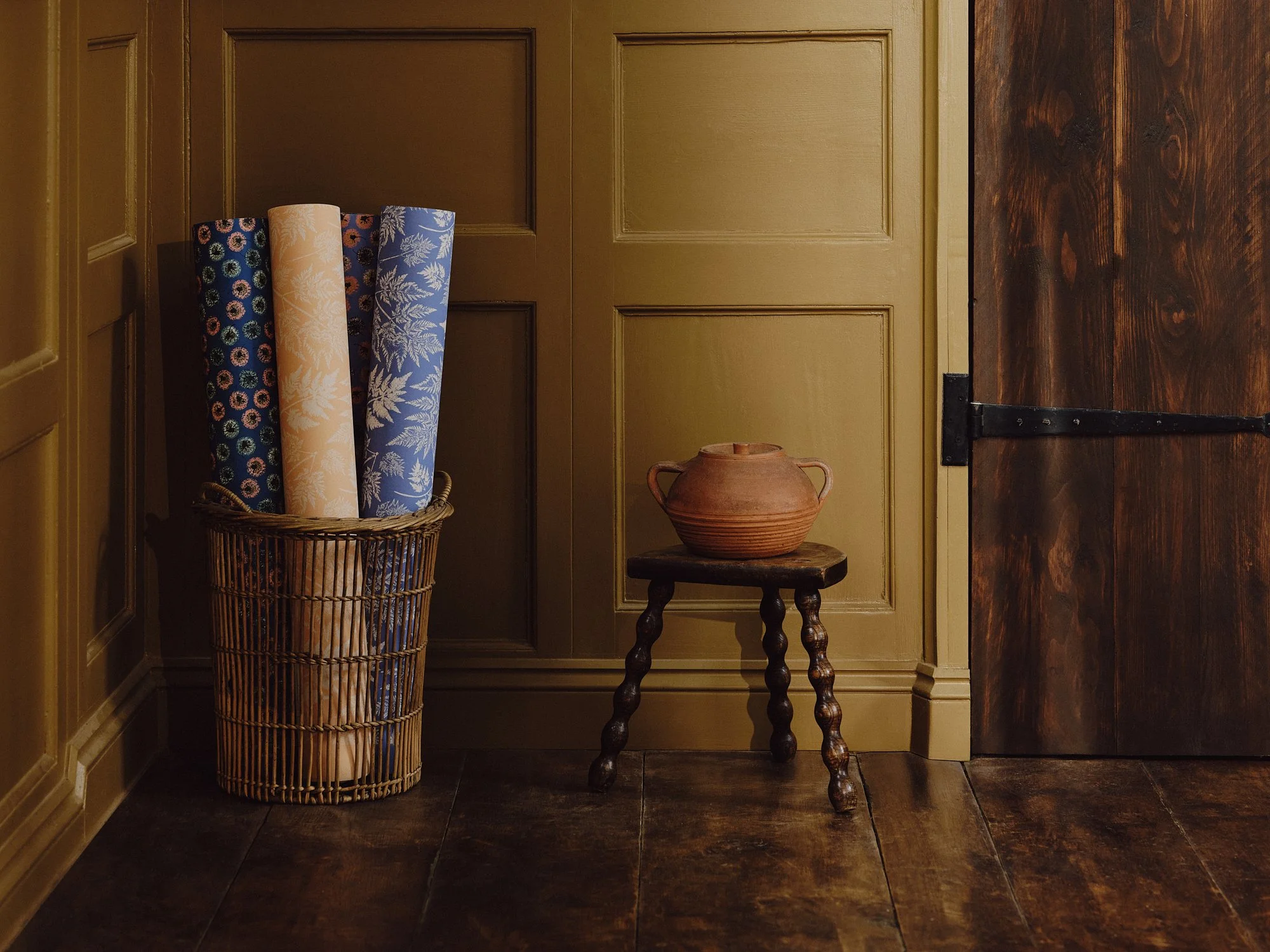 Basket with three rolled-up patterned papers, small wooden stool with a clay pot, wooden door and paneled wall in a room with wooden flooring.