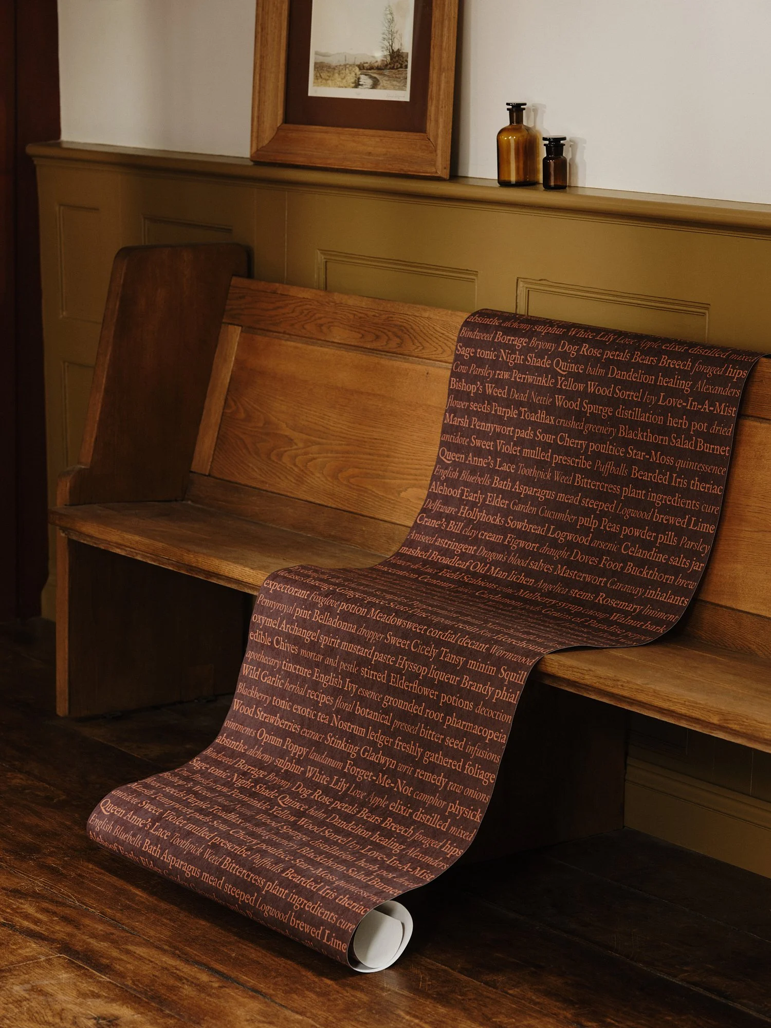 A wooden bench and a table with a brown and black printed paper scroll on a hardwood floor. The wall behind has a picture frame with a landscape photo and bottles on a ledge.