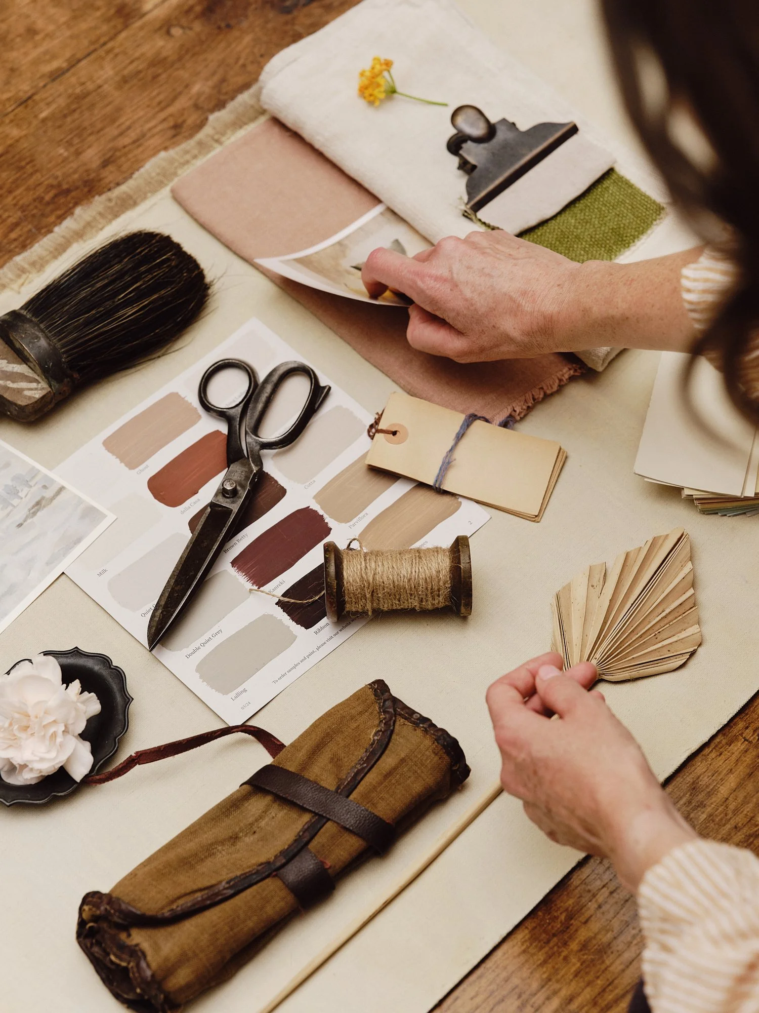 A person is working on a design project with fabric swatches, paint color samples, a paintbrush, scissors, thread, a paint sample fan deck, a small book, and decorative flowers on a table.