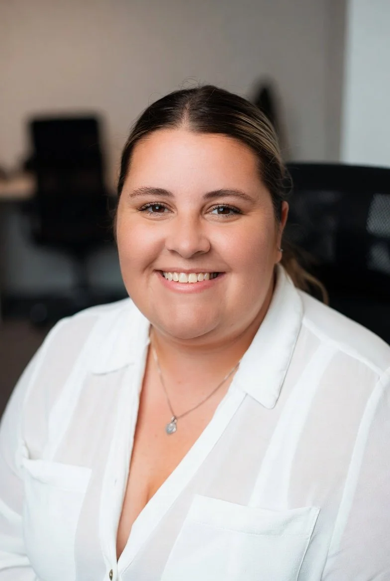 A smiling woman with brown hair tied back, wearing a white blouse and a necklace, sitting in an office chair.