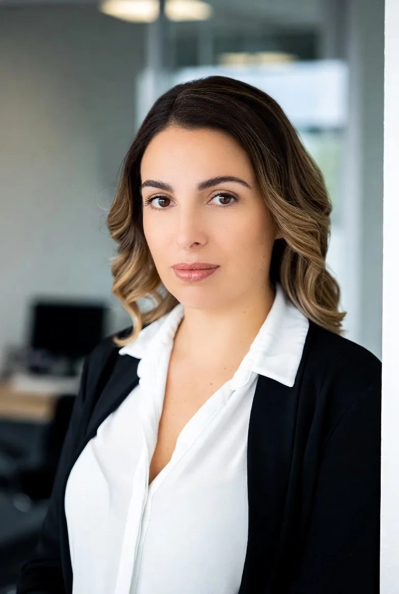 A woman with shoulder-length wavy brown hair, wearing a white blouse and black blazer, standing in an office setting.
