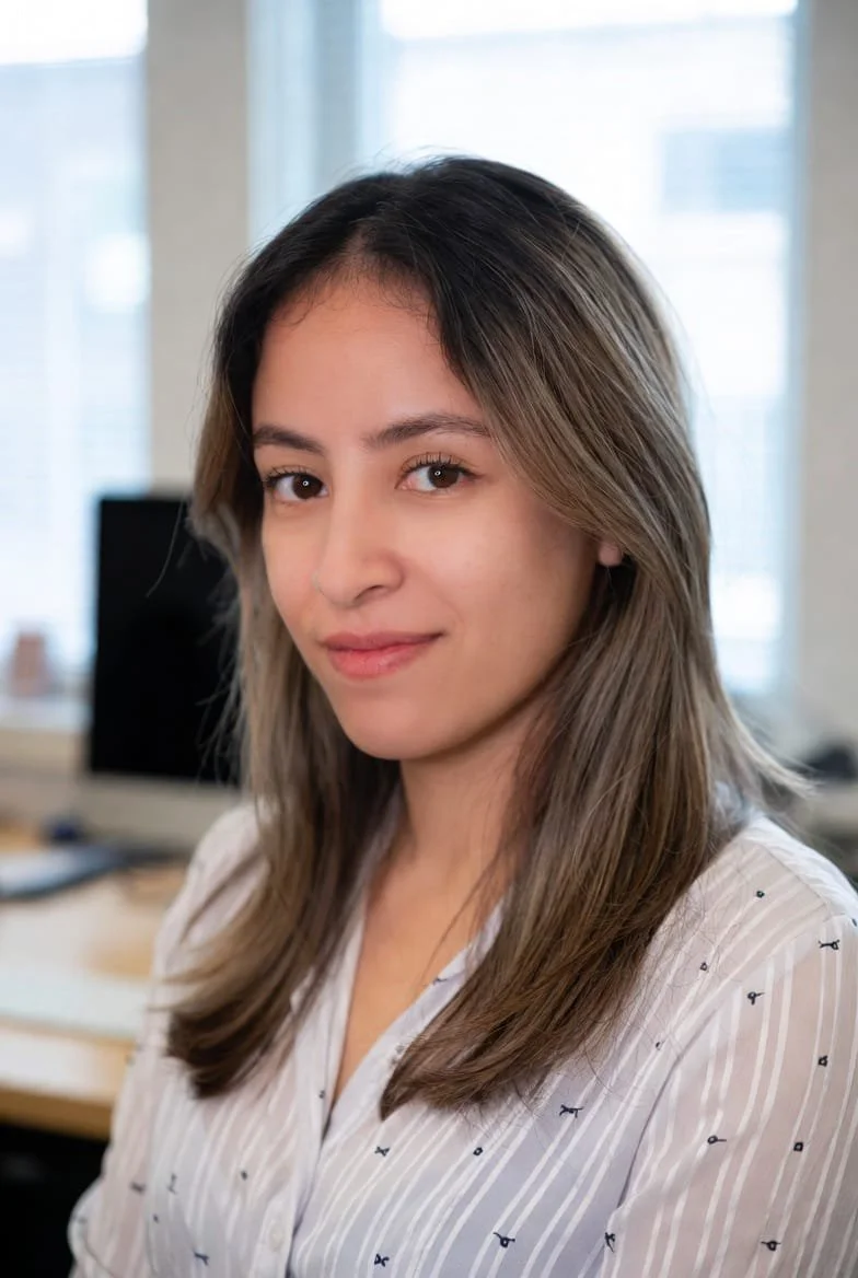 A young woman with light skin and shoulder-length brown hair with blonde highlights, wearing a white shirt with small dark patterns, sitting in an office with large windows and a computer monitor in the background.
