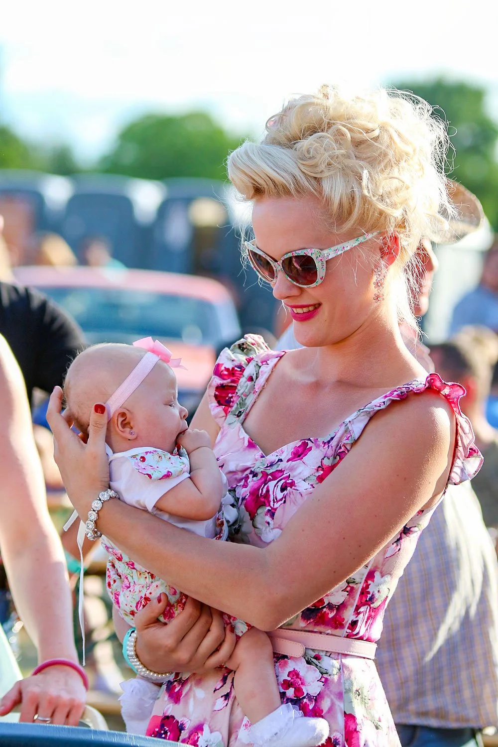 Une femme blonde portant des lunettes de soleil à motif floral tient un bébé dans ses bras lors d'un événement en plein air, avec des personnes et des voitures en arrière-plan.