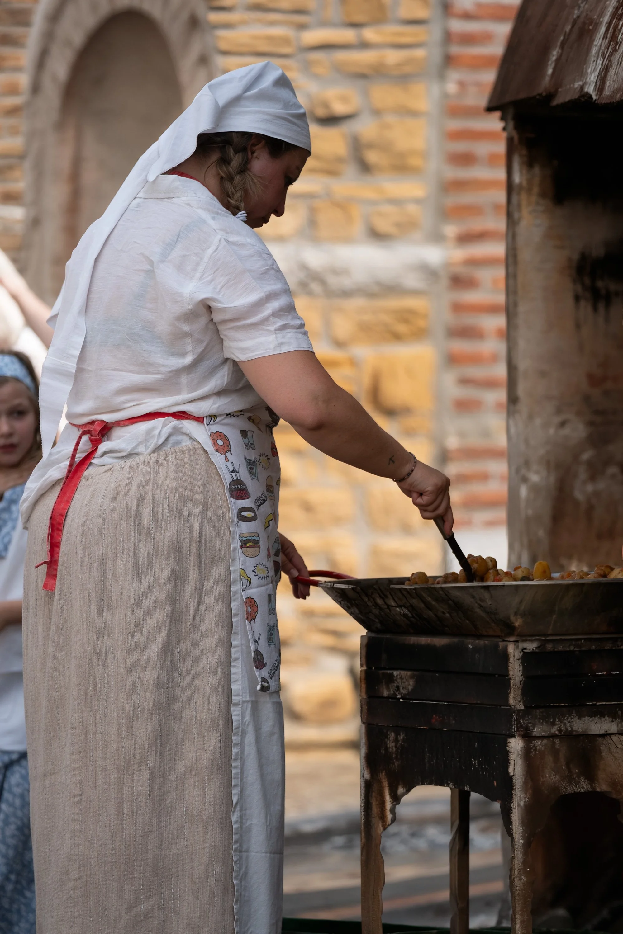 Une femme habillée en vieille mode, portant un bonnet blanc, cuisine des morceaux de pommes de terre sur un gril en pierre dans un environnement rustic, avec un mur en pierre en arrière-plan.