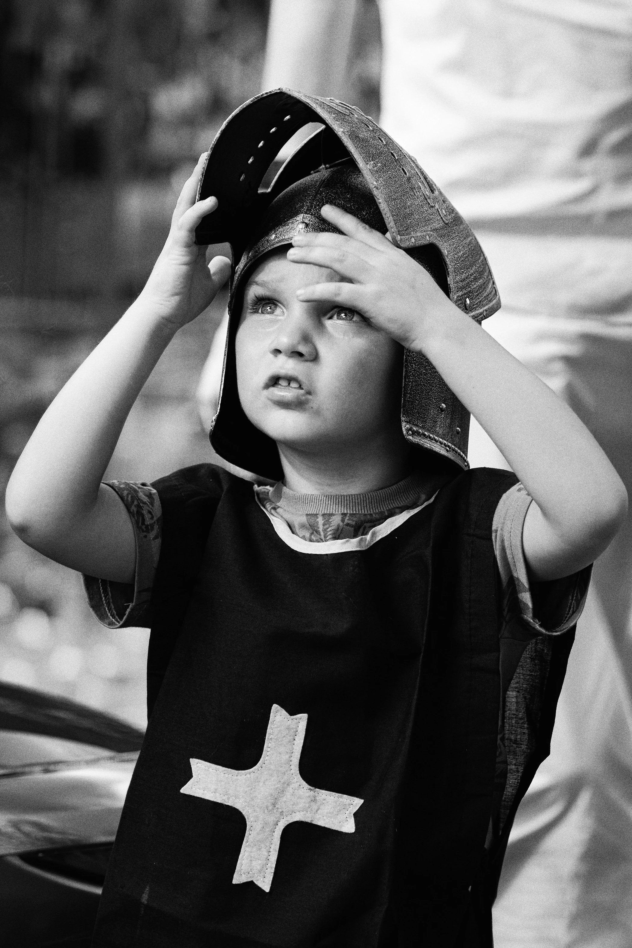 Jeune garçon portant un casque de pompier et un gilet avec une croix, regardant vers le haut, dans un style en noir et blanc.
