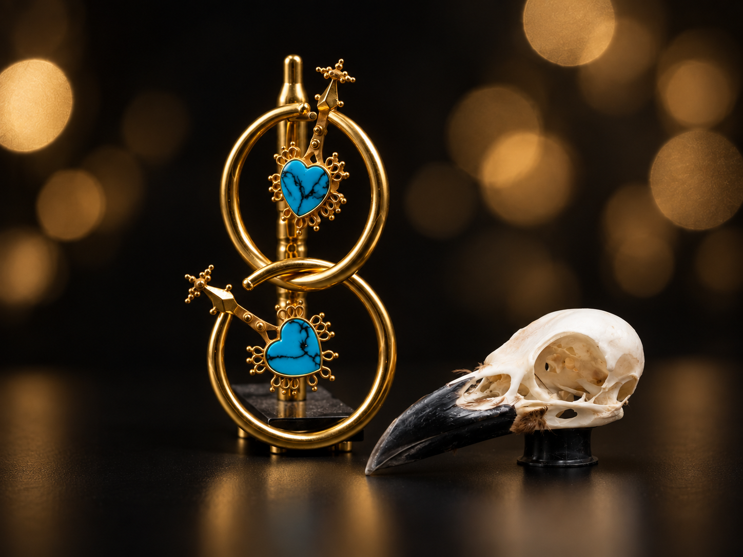 Boucles d'oreilles en or avec des coeurs en turquoise, et un petit crâne d'oiseau en os brillant avec un bec noir devant un fond noir à lumières dorées floues.