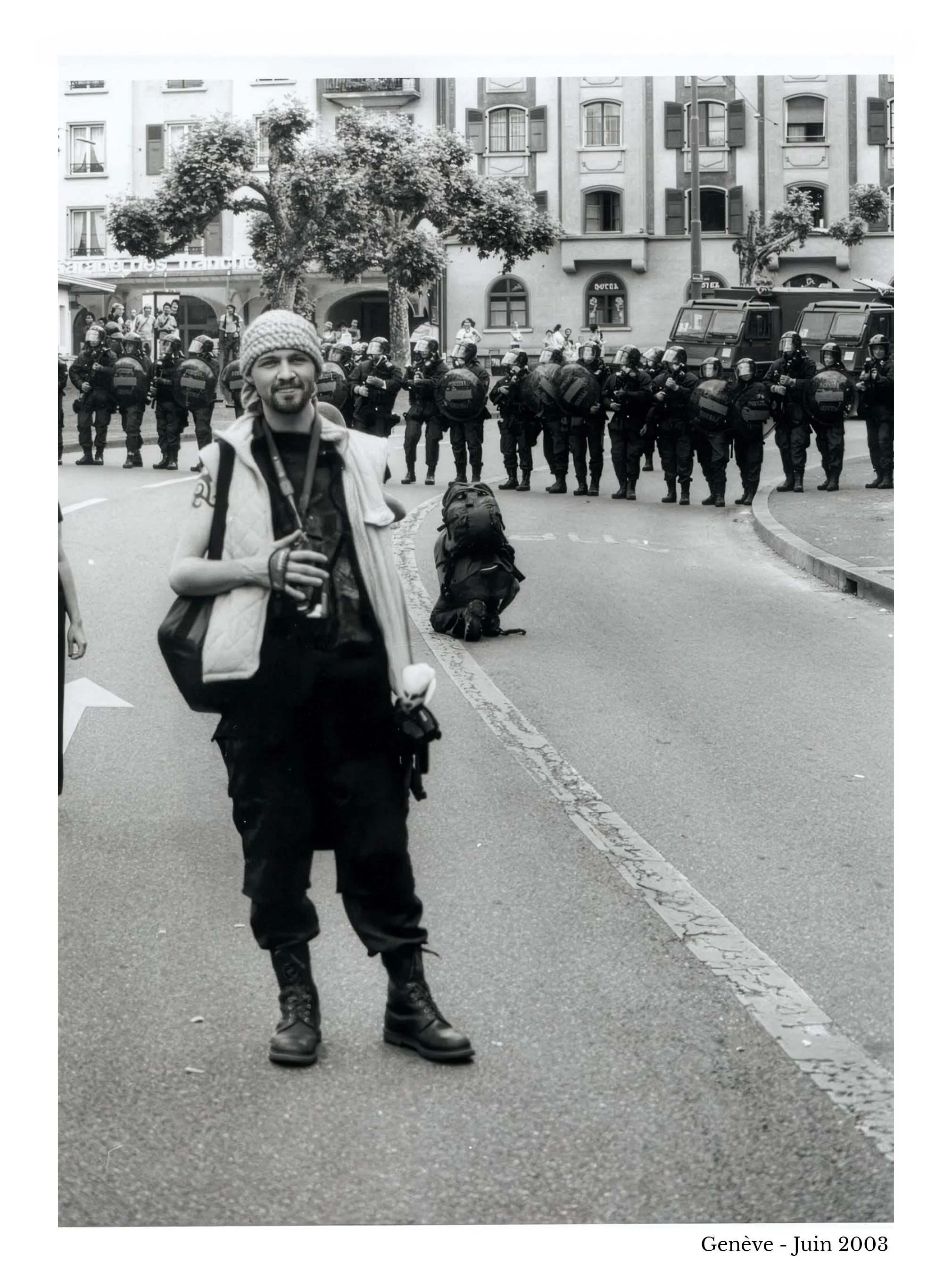 Photo en noir et blanc prise à Genève en juin 2003 montrant Raphaël Haentjens avec un sac à dos et une caméra, debout sur la route, avec une ligne de policiers en formation en arrière-plan et une personne agenouillée sur le sol.