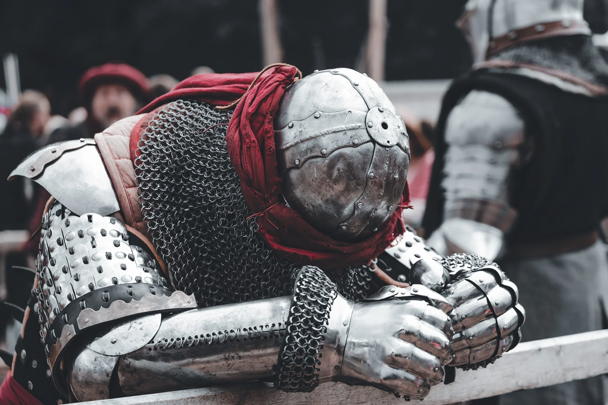 Armure médiévale en métal avec des mailles en acier, un casque en acier, et un tissu rouge autour du cou, avec des personnes en arrière-plan.