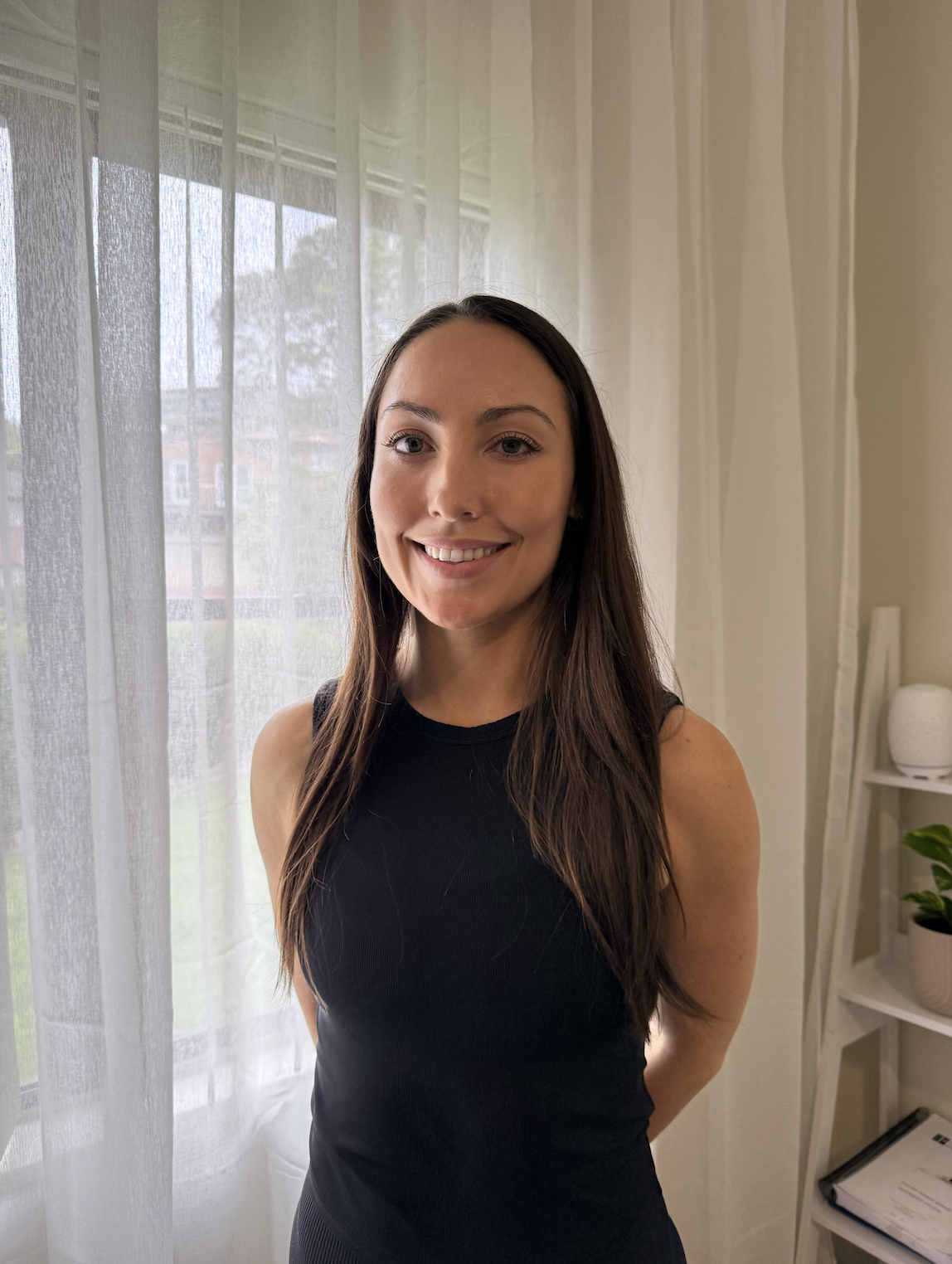 A woman with long brown hair wearing a sleeveless black top, standing in her Pilates studio near a window with sheer white curtains, with a bookshelf and potted plant in the background.