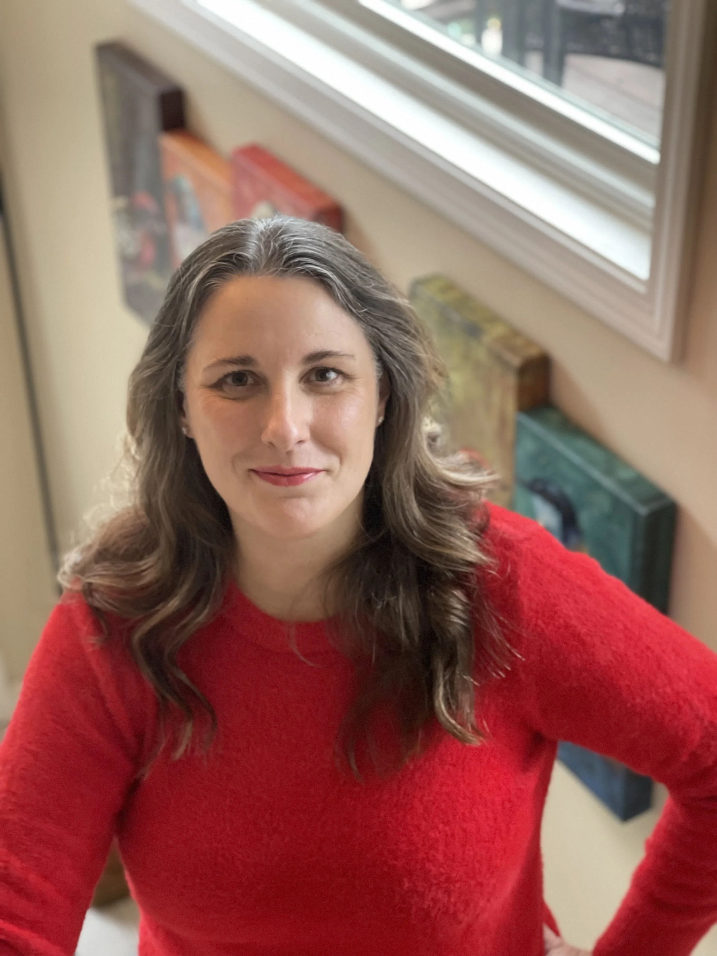 A woman with long, wavy brown hair wearing a red sweater, standing indoors near a window with bookshelves in the background.