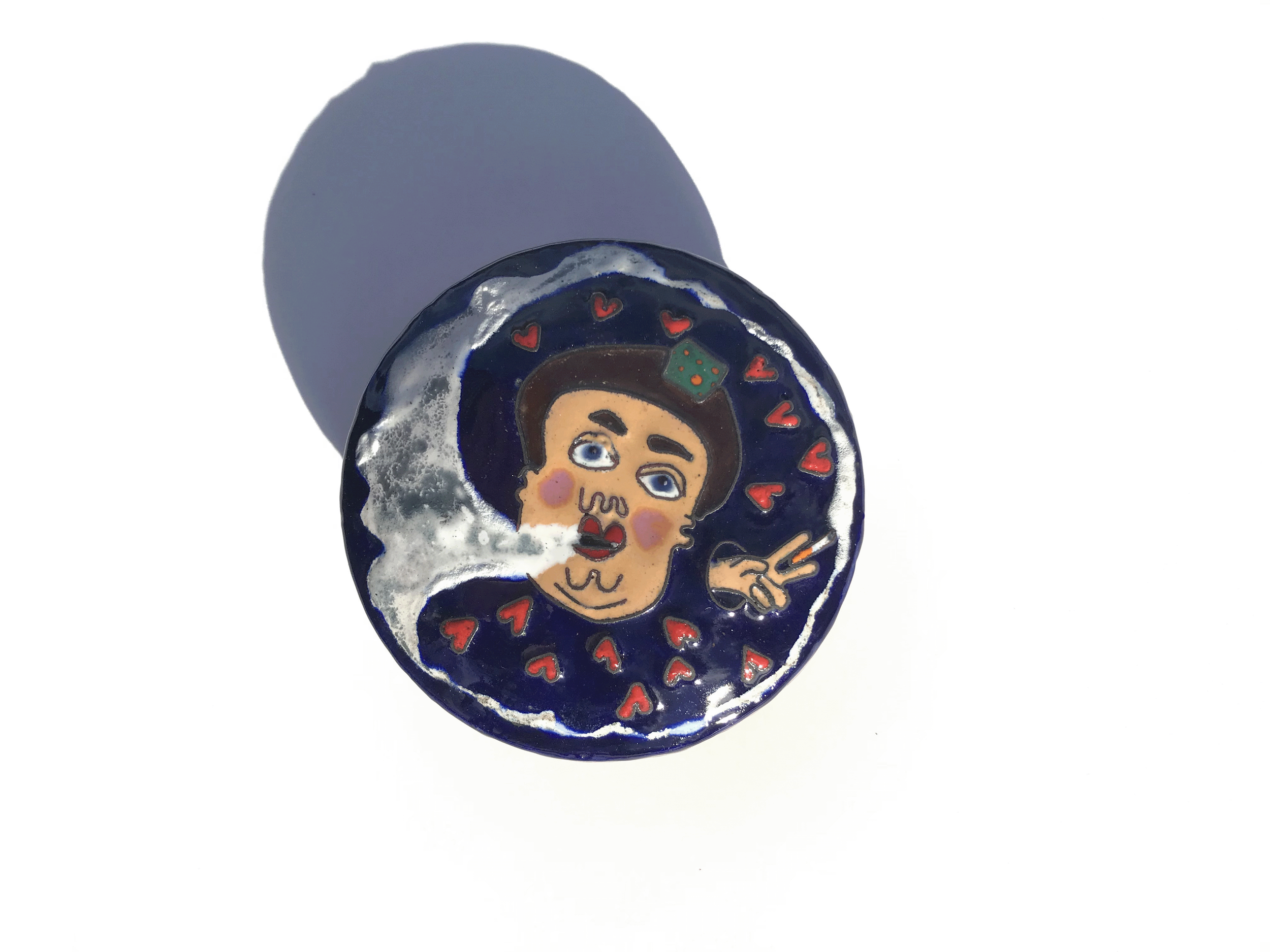 En rund smykkeæske i kobber med emaljedekoration, som forestiller en person, som som ryger, og røgen omringer ansigtet. Baggrunden er mørkeblå.