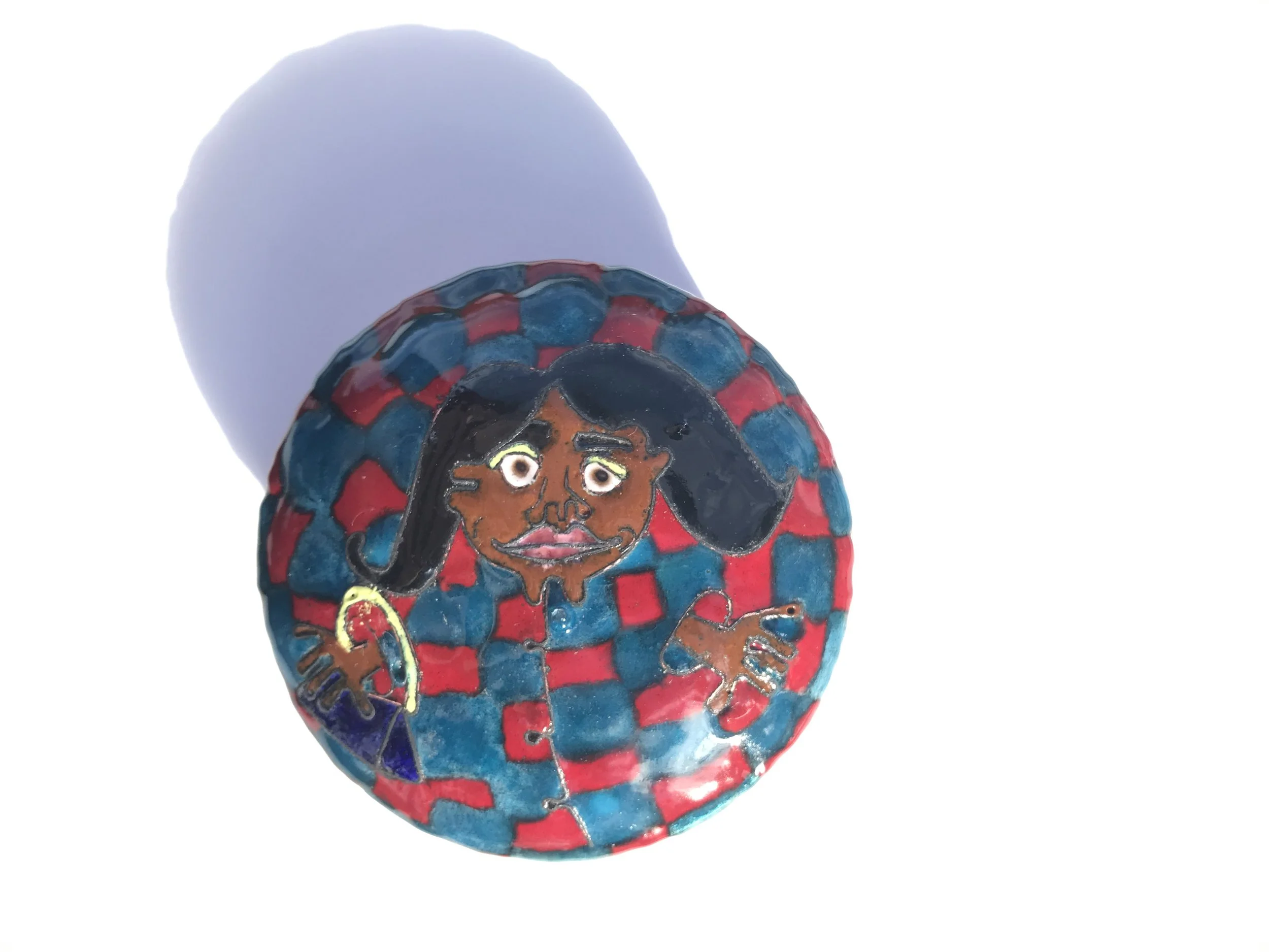En rund smykkeæske i kobber med emaljedekoration, som forestiller en person, som har mørkt hår, stort smil, og holder en taske. Baggrunden er en rød og blå ternet mønster. 