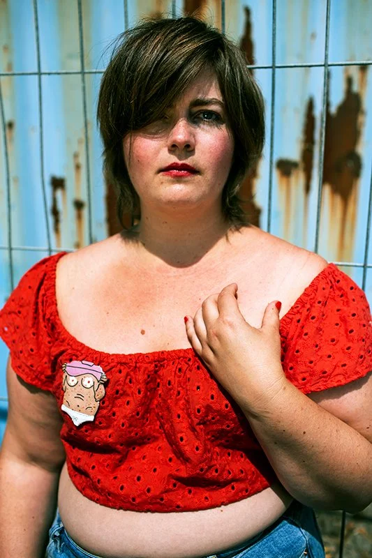 Ida Rud iført en rød top med FEMME AND FAB briche, som står foran en rusten metalvæg.