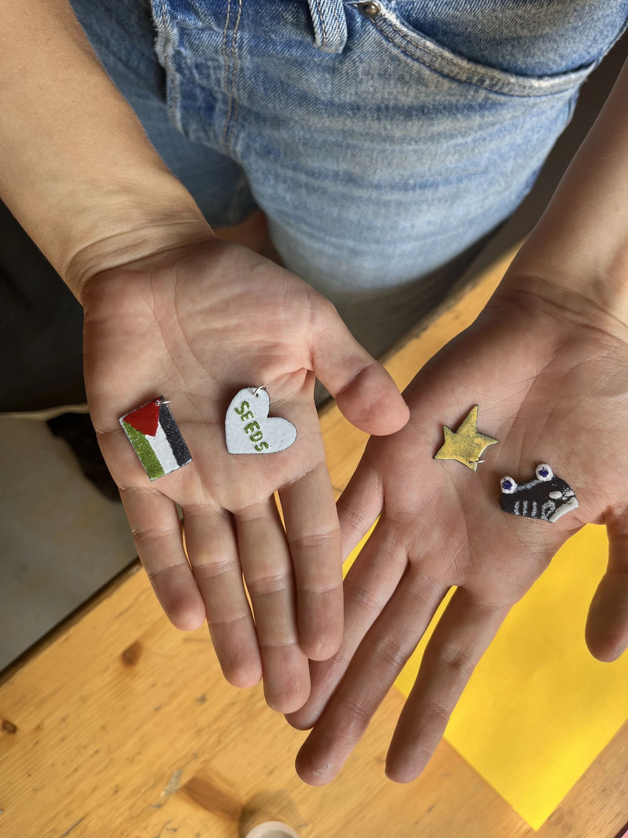 En person holder to hænder med fire små, farverige emaljevedhæng frem, der ser ud til at være små figurer. Billedet er to af personens hænder.