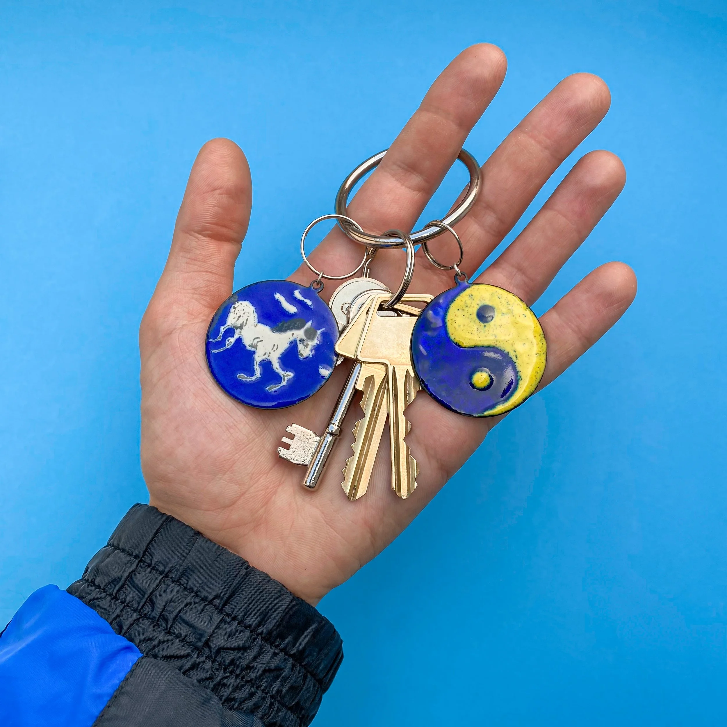 Hånd holder nøgler med yin-yang symbol i emalje, en hest med blåt baggrund i emalje op mod en blå baggrund.
