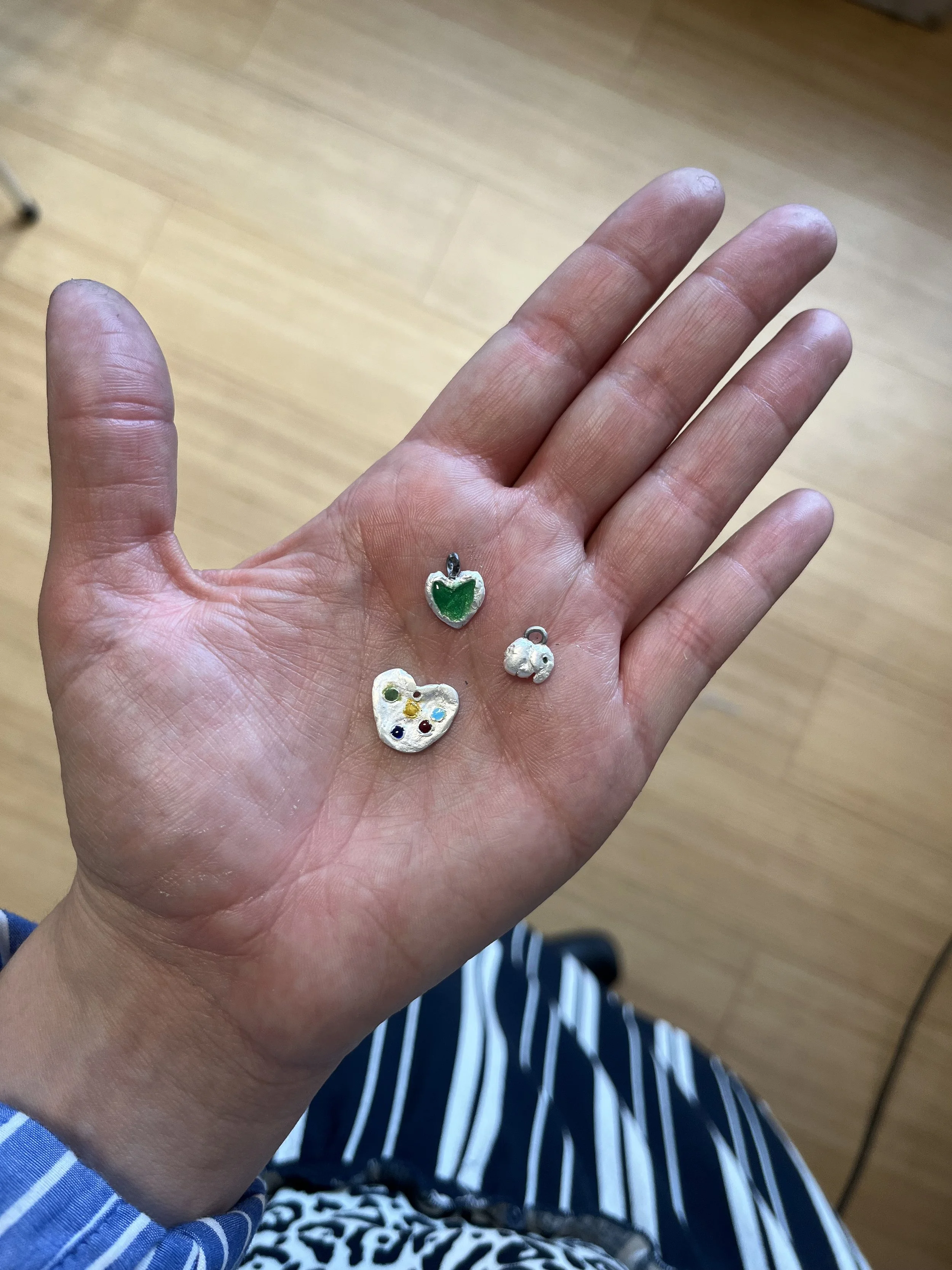Hånd holder tre små charms, en med grøn emalje, et sølv hjerte med farvede prikker, og en lille sød elefant.