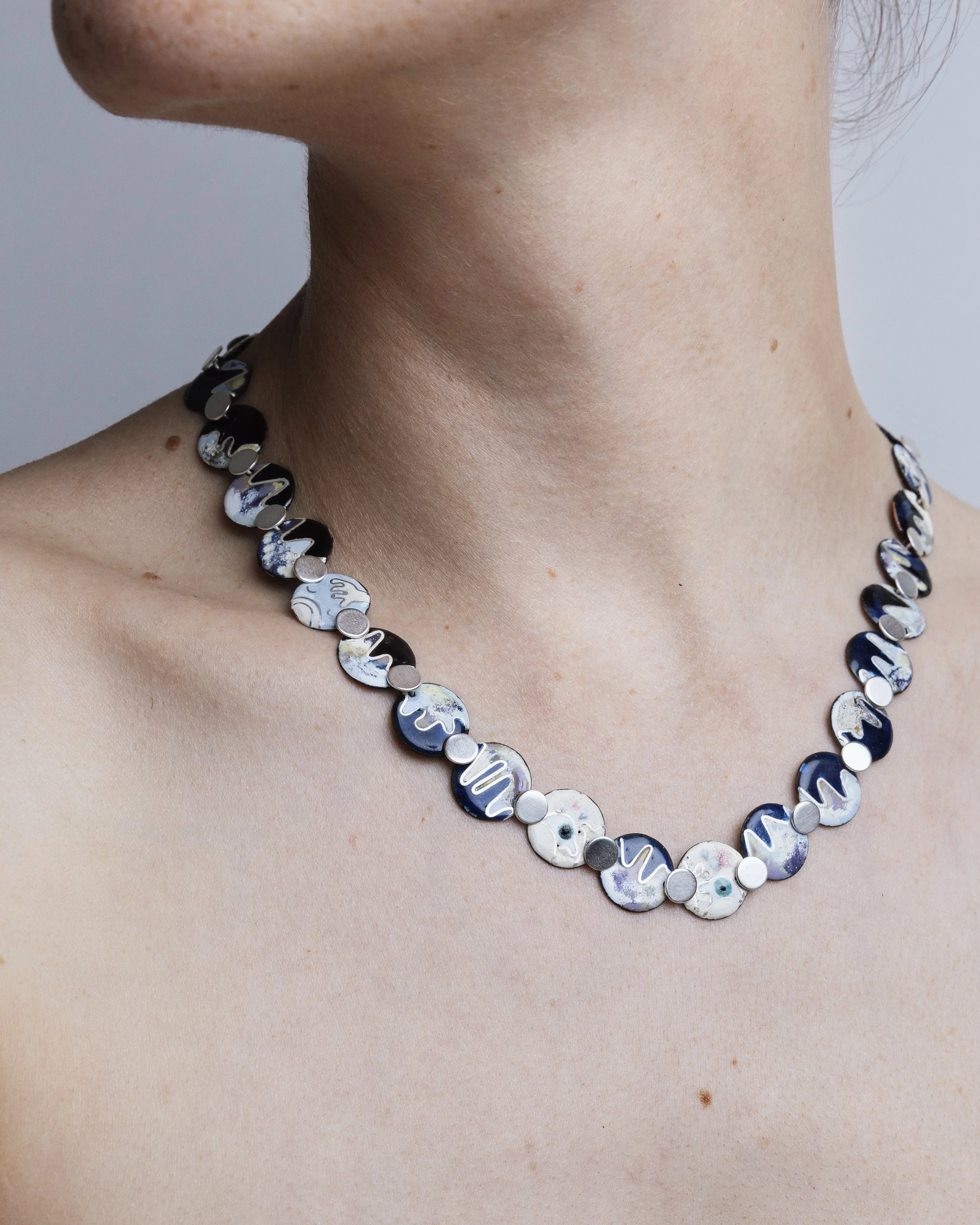 Nærbillede af en kvindes hals med en halskæde lavet af cirkulære emaljevedhæng i forskellige farver og mønstre.