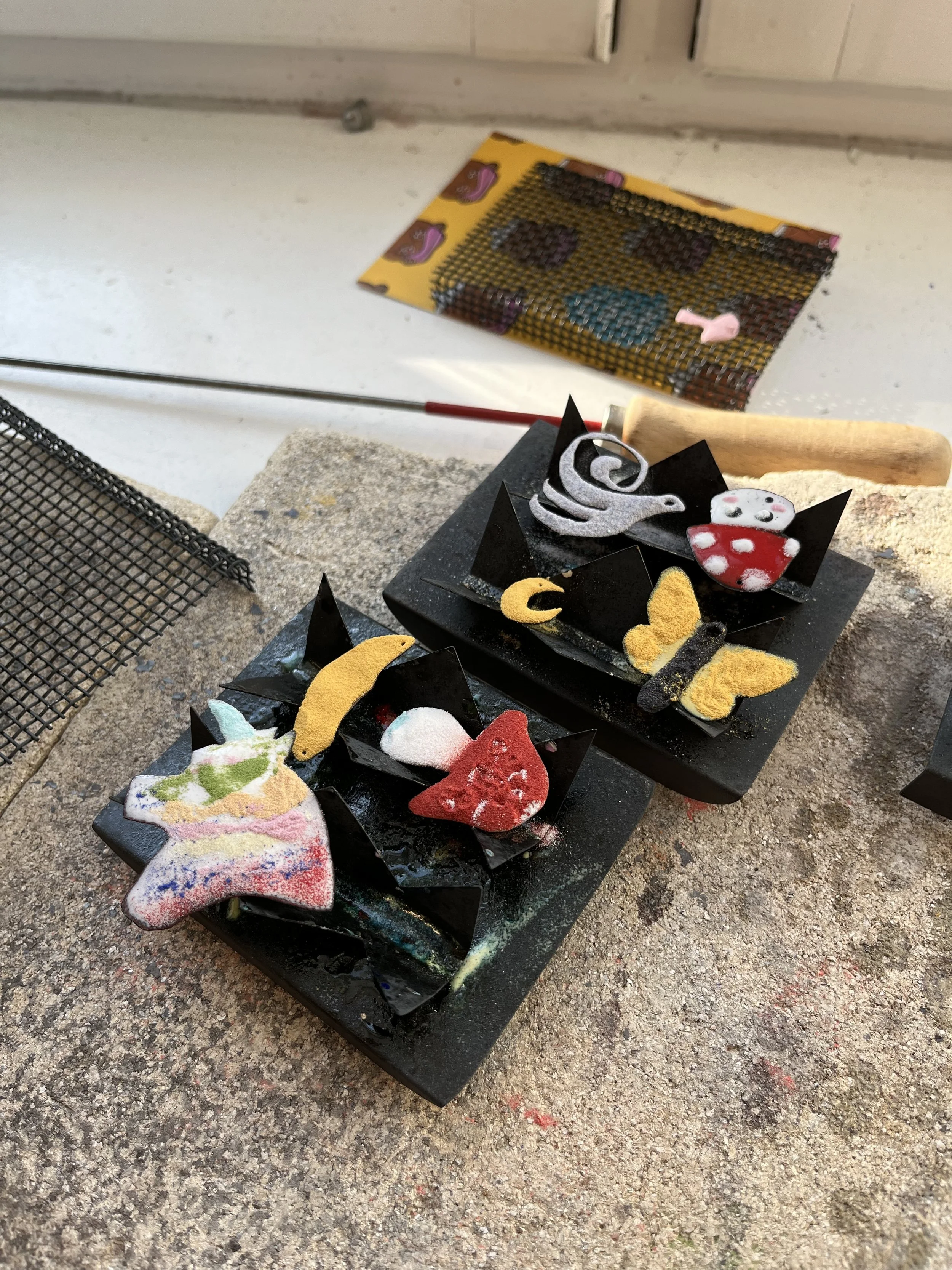 Forskellige farverige korsstingsbroderede figurer og emojier, inklusive en hjerte, stjerne, måne, sommerfugl og fyr. Nogle af figurerne er lavet af stof og har glitter.