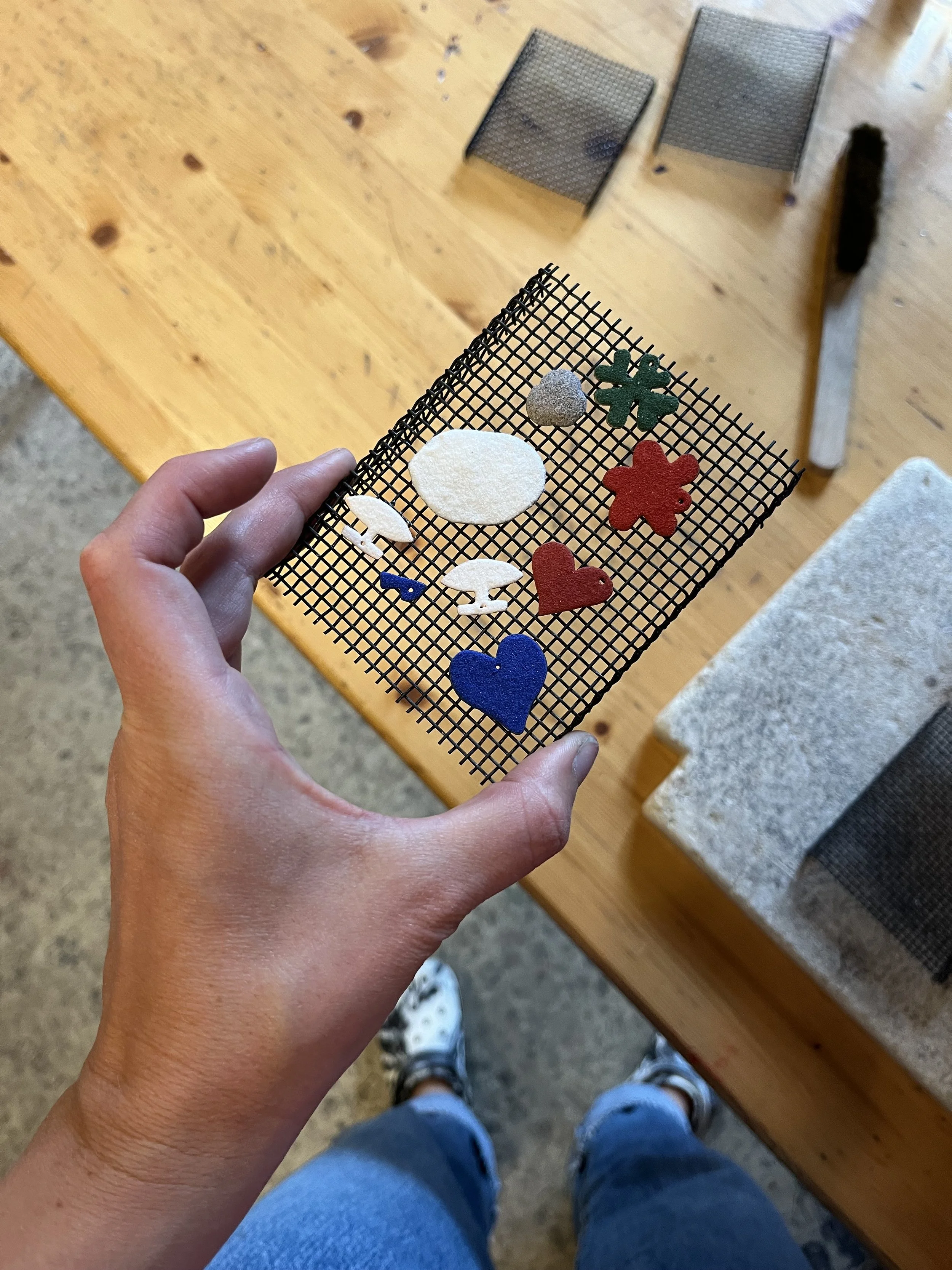 Hånd holder en loddenet med forskellige emalje figurer i farverne grøn, rød, blå, og hvid, som er lavet til en sav selv dit eget design workshop.