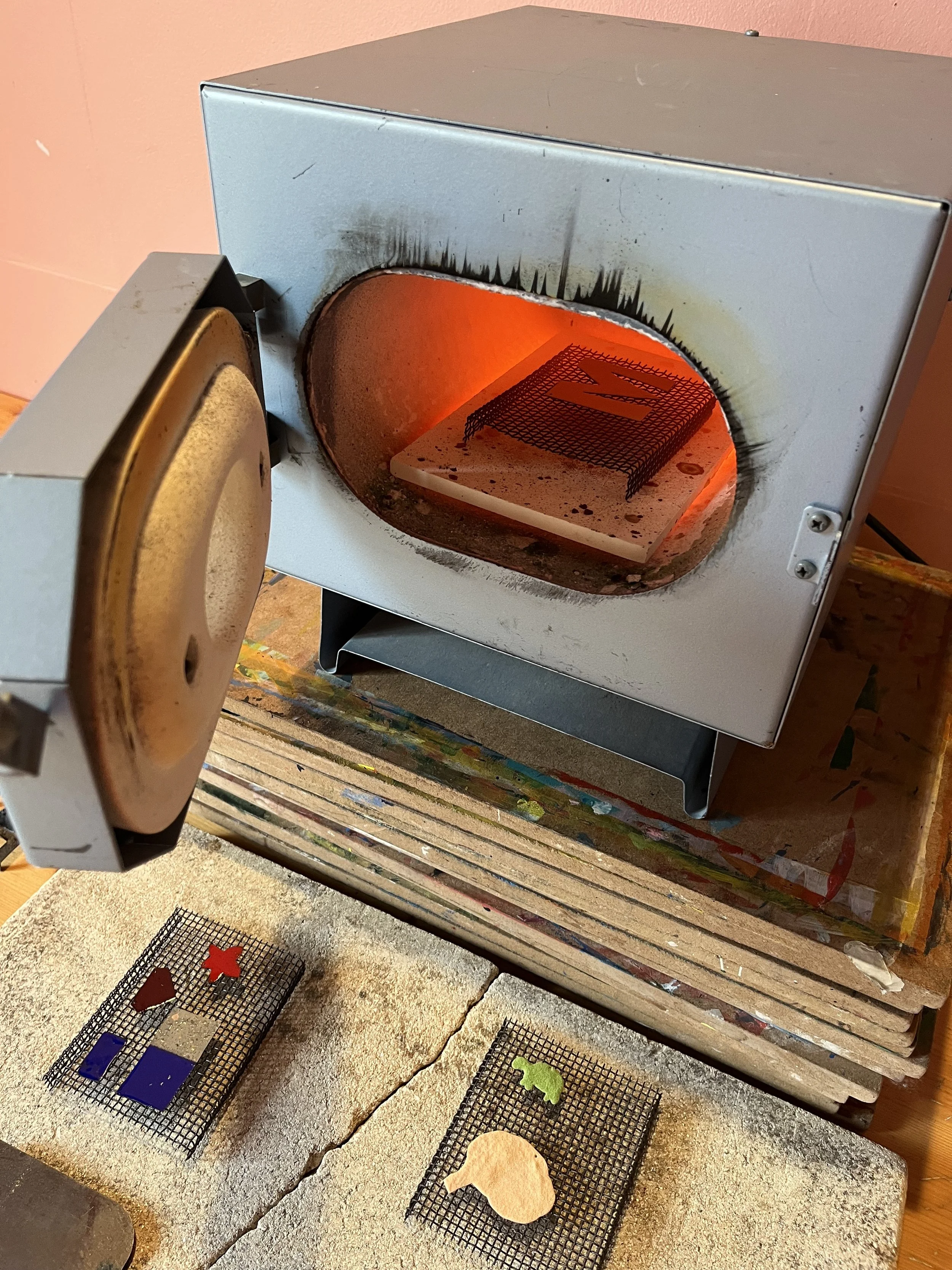 En glasovn, der bruges til at brænde eller smelte små metal eller glaspålæg og nogle farvede metal- eller glasfigurer, er placeret på en brændovn. Ovnen er tændt, og der er registrerede figurer foran ovnen.