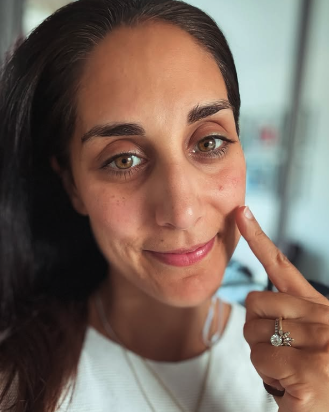 Close-up of Dr Sara Kayat with brown hair, hazel eyes, and light skin, smiling and touching her cheek with her finger, wearing a white top and a diamond ring.
