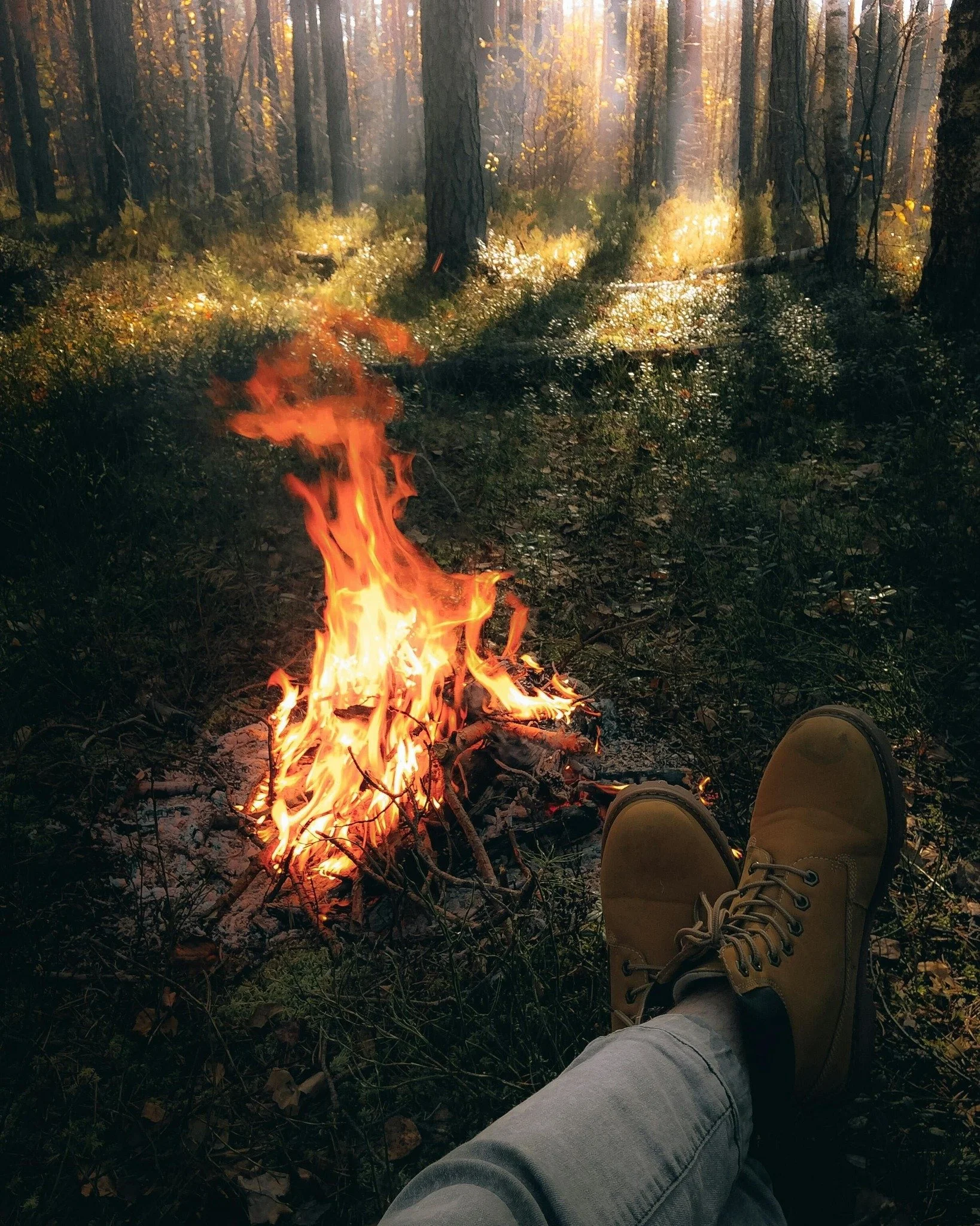 Person relaxing in a forest, lying back with legs extended, wearing yellow boots and gray pants, beside a campfire.