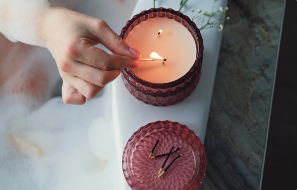 A hand is using a match to light a candle in a glass jar with a textured red exterior, placed on a white surface next to a matching red lid and a small vase with green flowers.